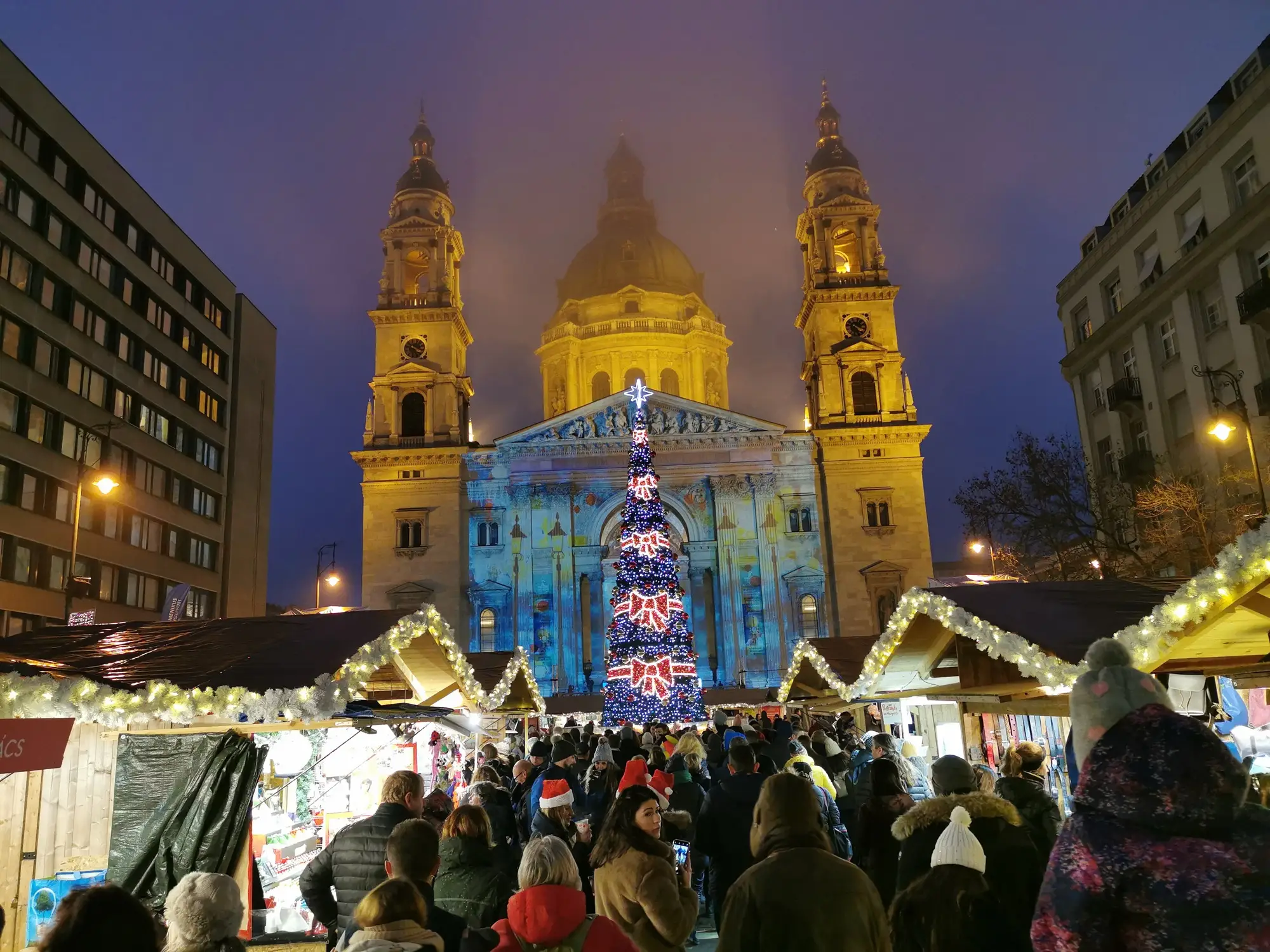 St Stephen's Basilica lit with a festive light projection behind a tall Christmas tree and the wooden stalls of the Advent Bazilika market at dusk