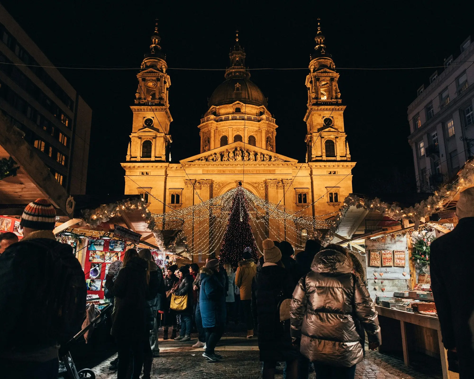 St Stephen's Basilica floodlit at night above the wooden stalls of the Advent Bazilika market, with the lit Christmas tree and a crowd in front of the church