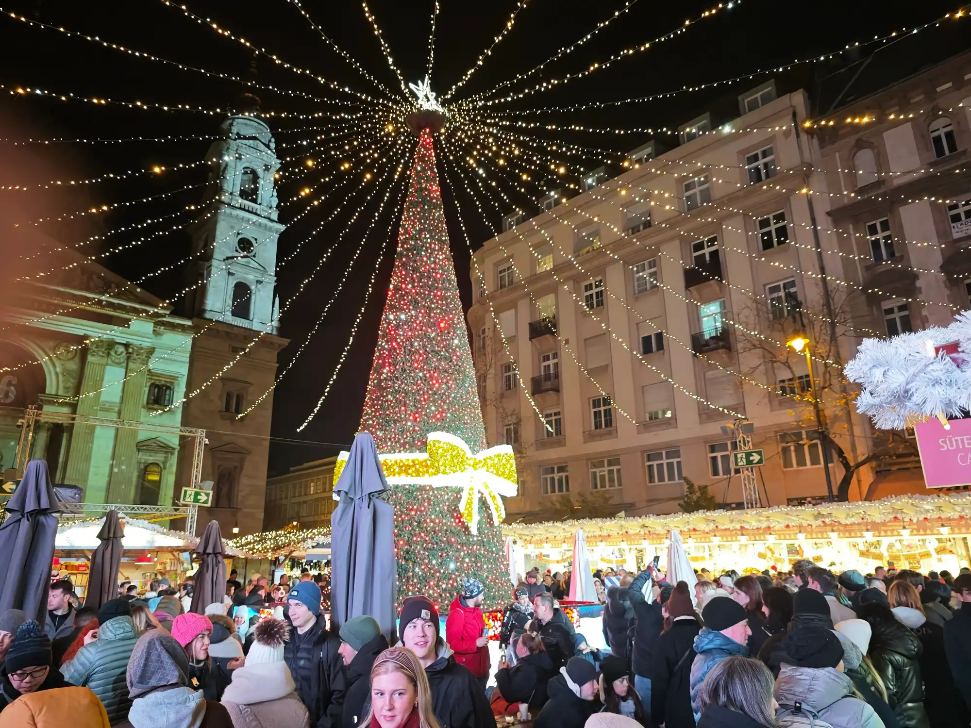 The giant Christmas tree at the Advent Bazilika market under a canopy of string lights, with a tower of St Stephen's Basilica on the left