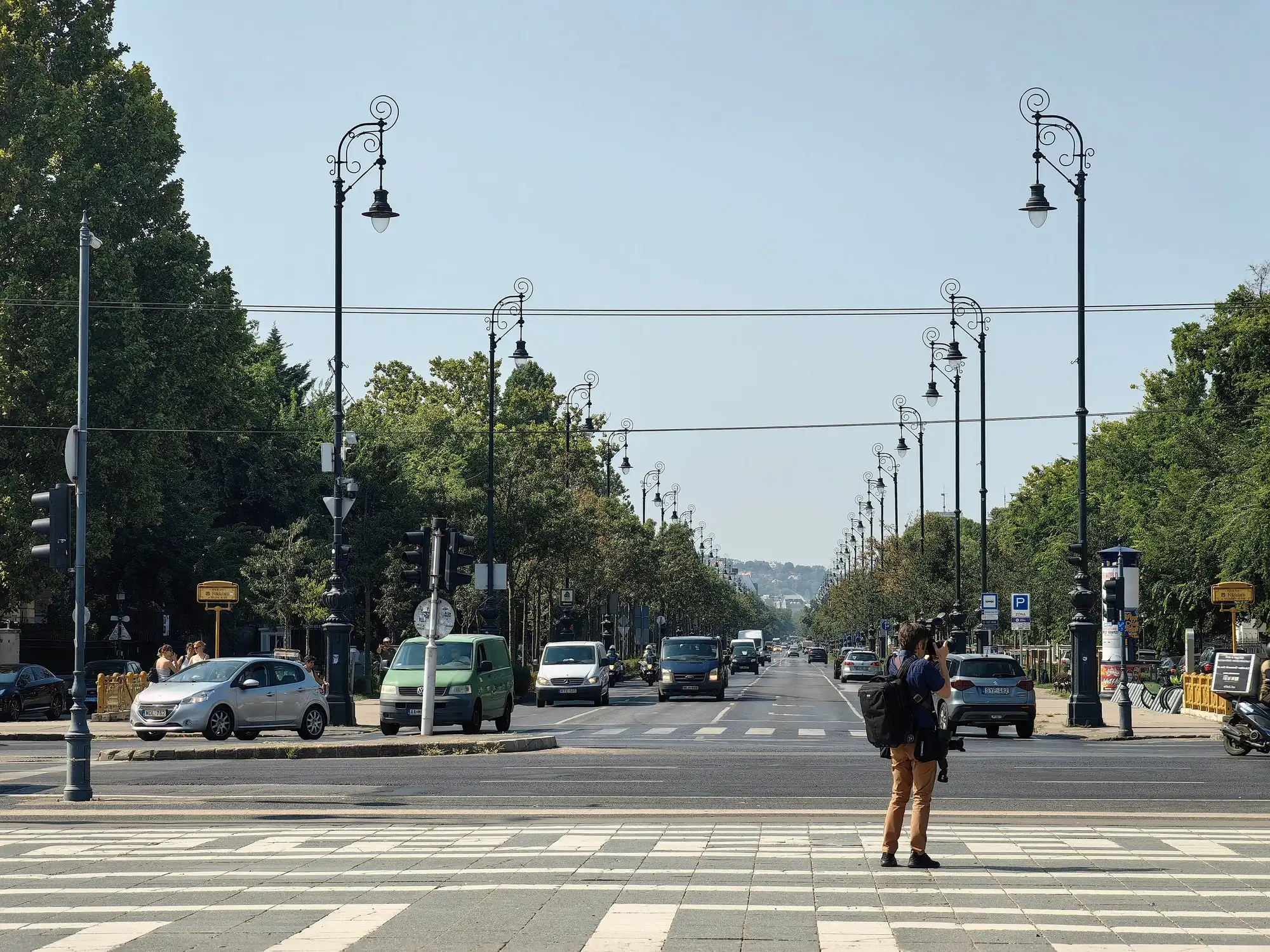 Andrássy Avenue at pavement level in Budapest, its lamp standards receding down a wide four-lane road with a pedestrian crossing in the foreground