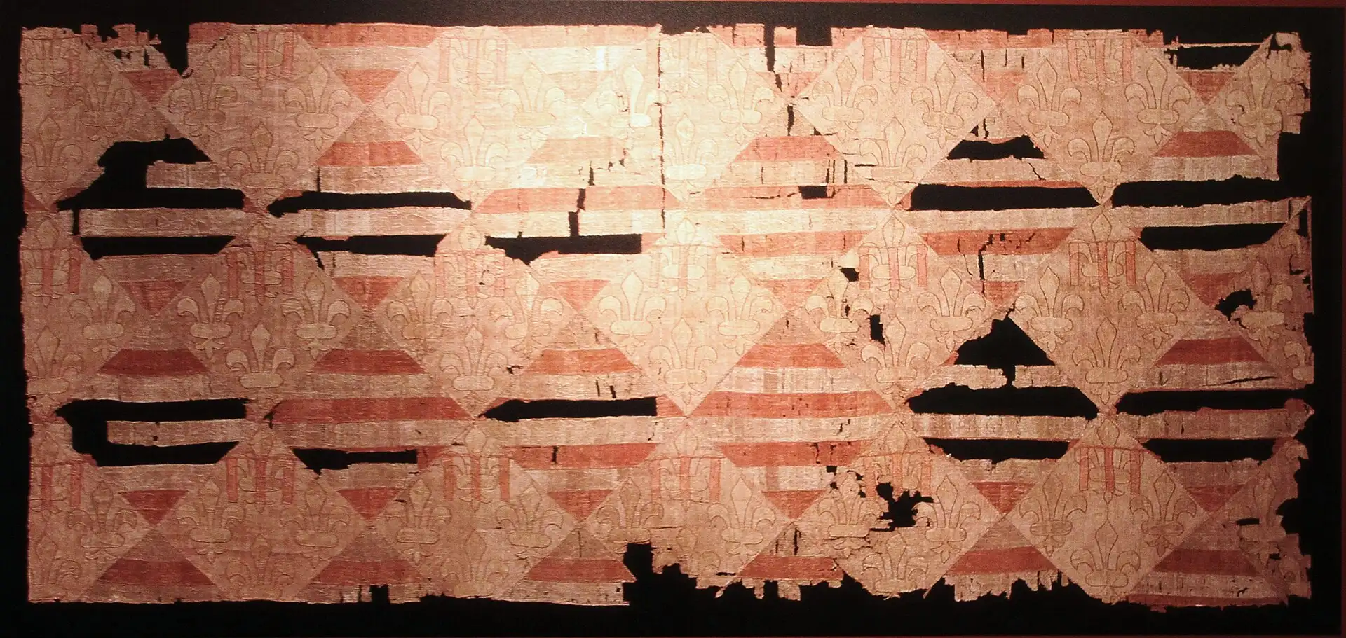 The Hungarian-Angevin silk hanging in its case at the Budapest History Museum, woven with fleurs-de-lis and the red and silver bars of the Árpáds in lozenge shields