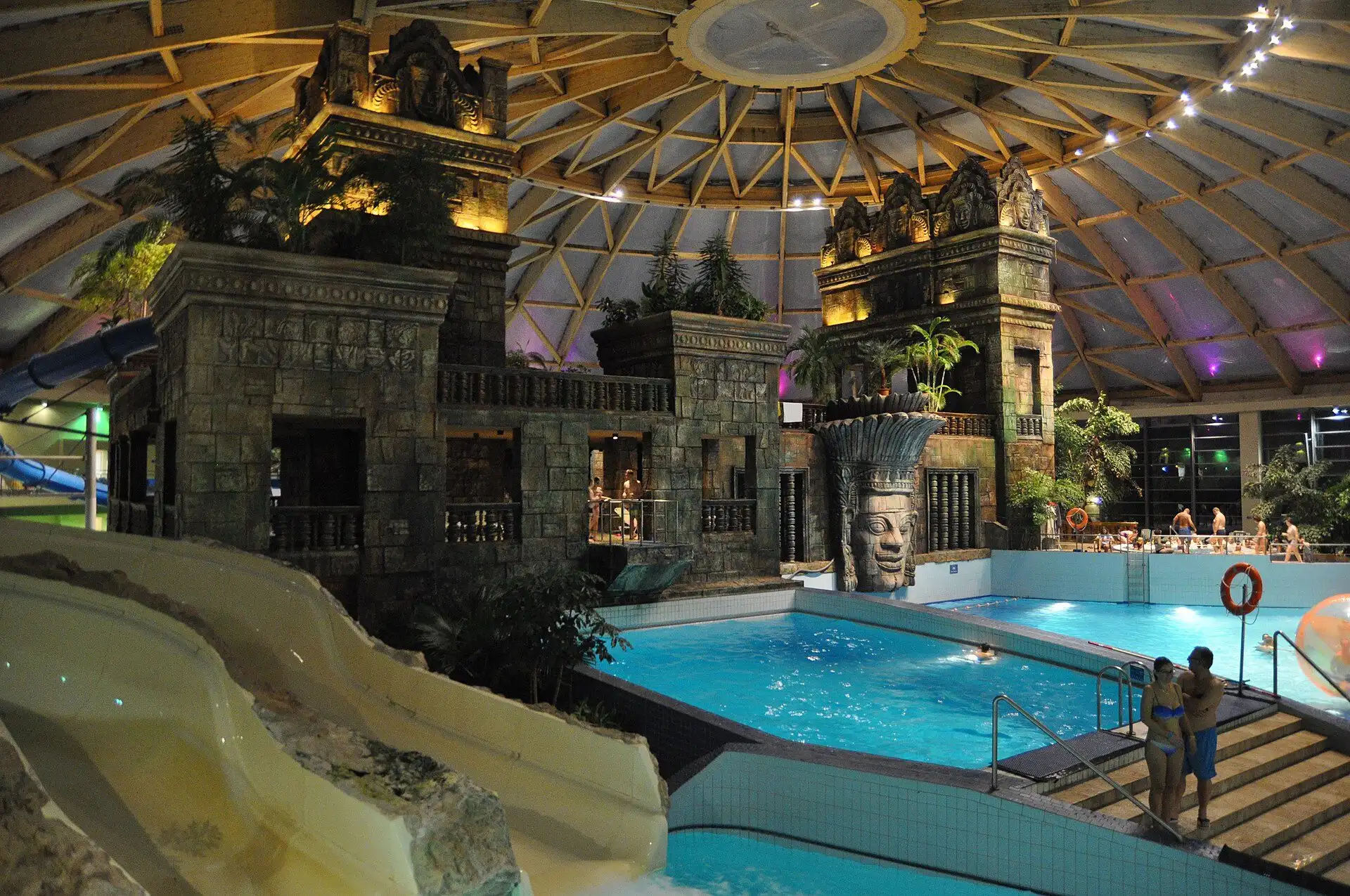 A slide run-out beside the indoor adventure pool at Aquaworld in the evening, with the temple lit up behind