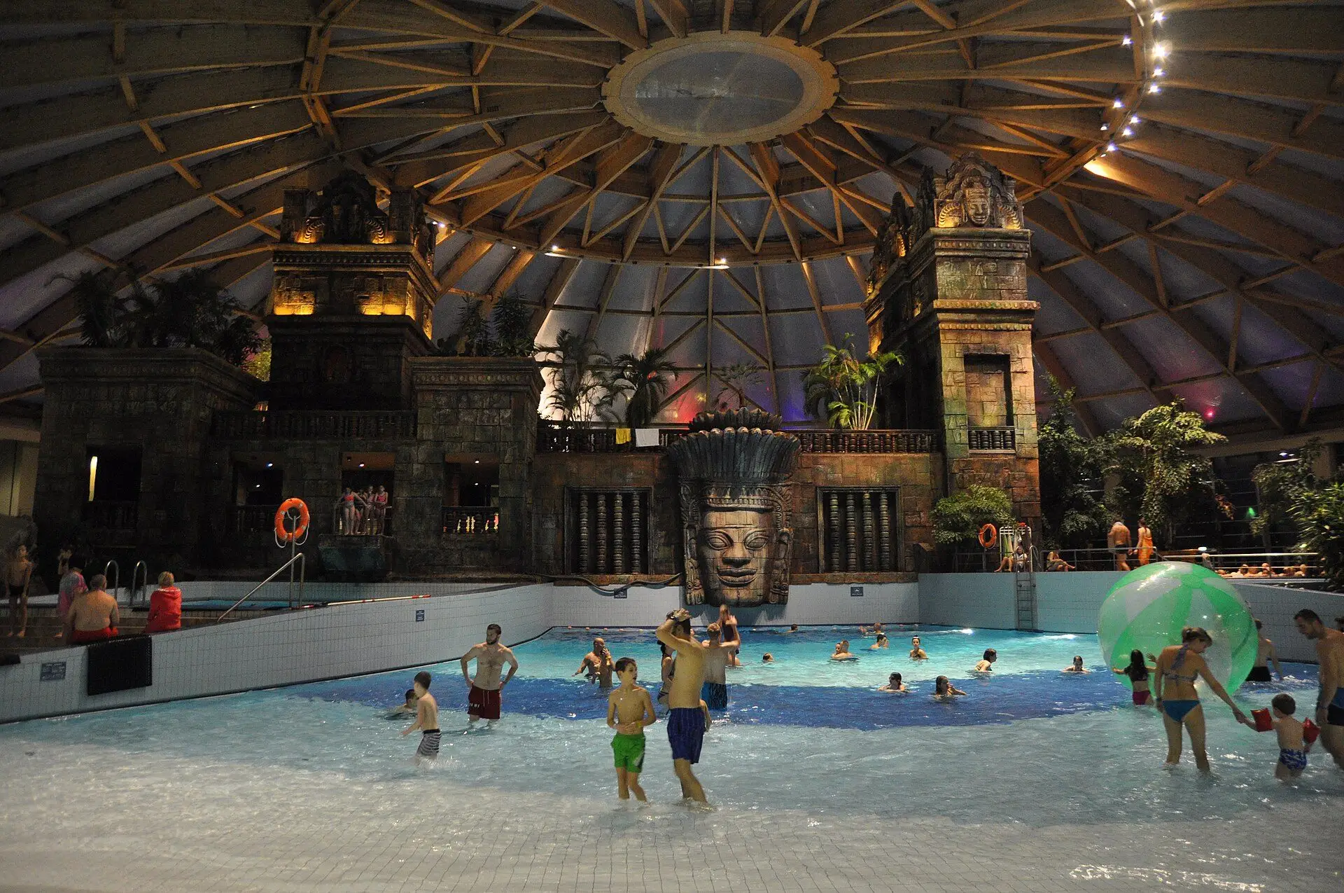 Children in the shallow end of Aquaworld's wave pool in the evening, a water walking ball on the water and a carved stone face on the temple behind