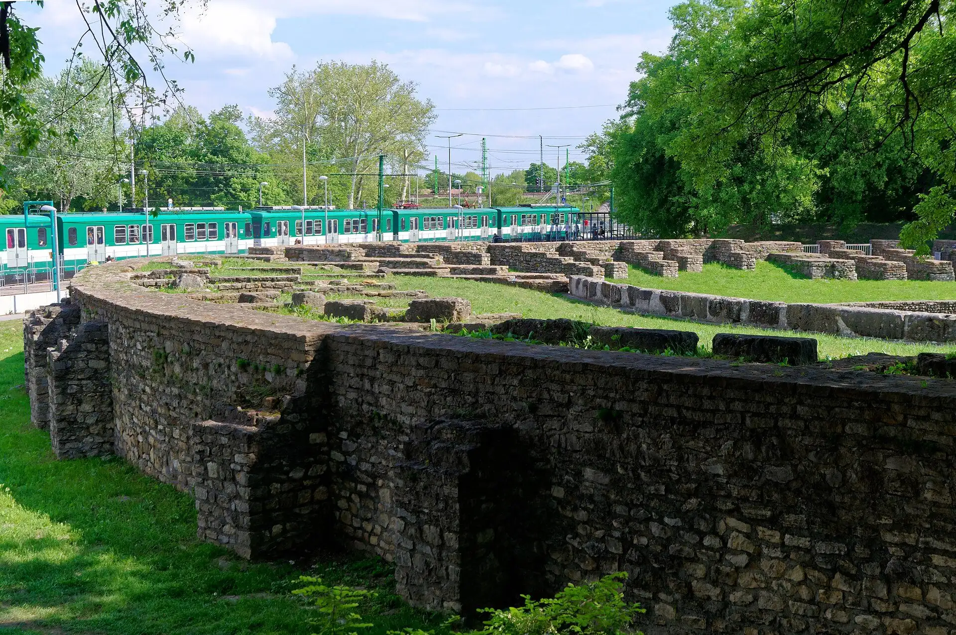 The curved stone ring wall and grass arena of the Aquincum civil amphitheatre, with a green suburban railway train passing behind it
