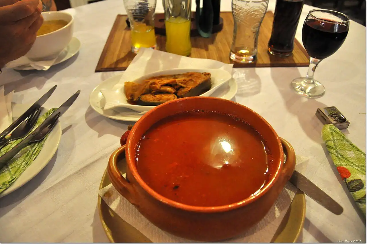 A clay pot of red Baja fish soup on a restaurant table, with a plate of fried fish and a glass of red wine behind it