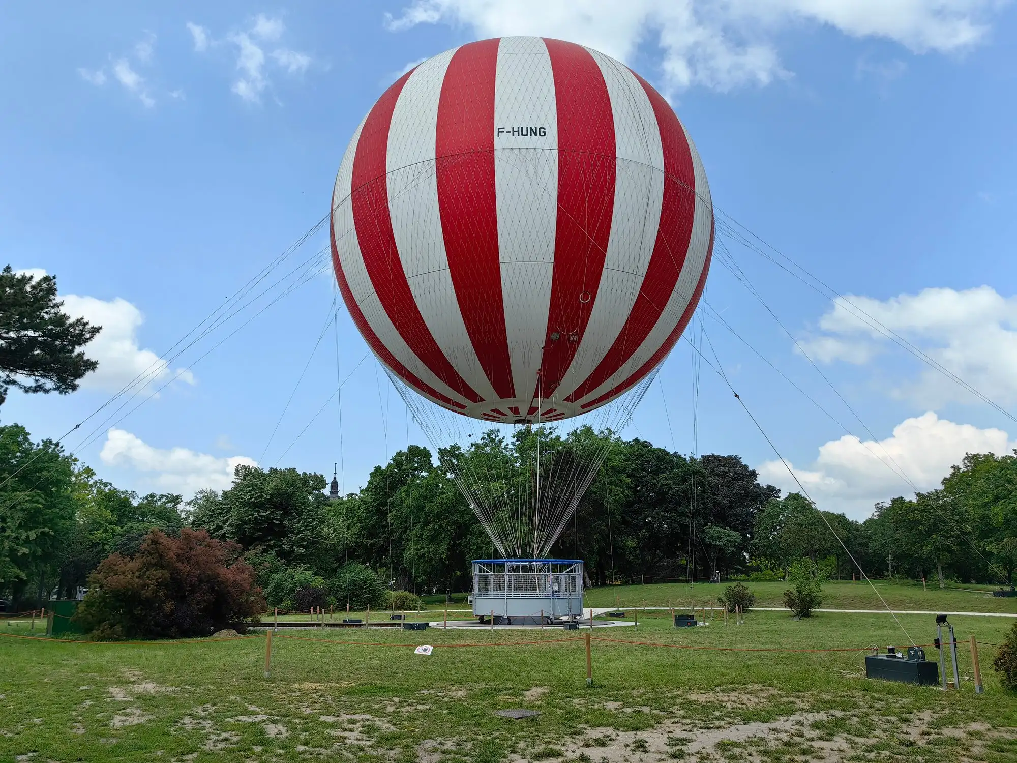 The red and white striped BalloonFly tethered balloon standing on its launch field on Mimosa Hill in Budapest's City Park under a blue sky