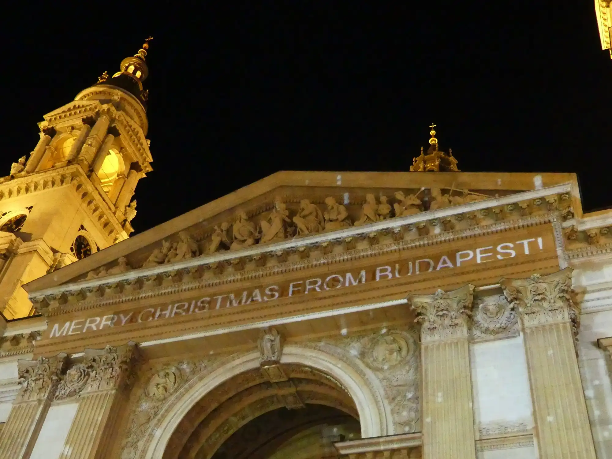 The front of St Stephen's Basilica at night with the words Merry Christmas from Budapest projected across the frieze and snowflakes of light on the stone