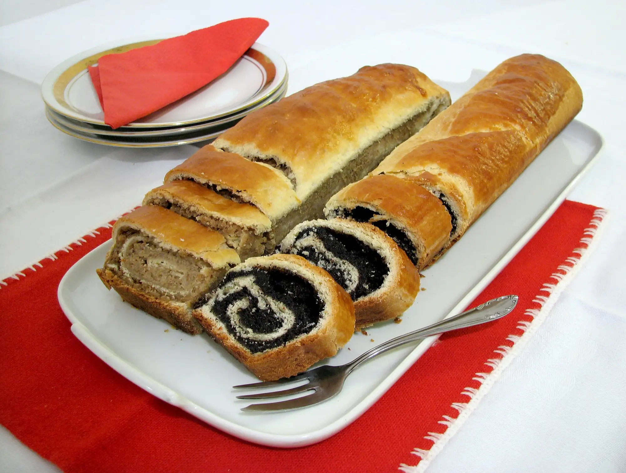 Two bejgli rolls, one walnut and one poppy seed, sliced on a white serving plate on a red cloth