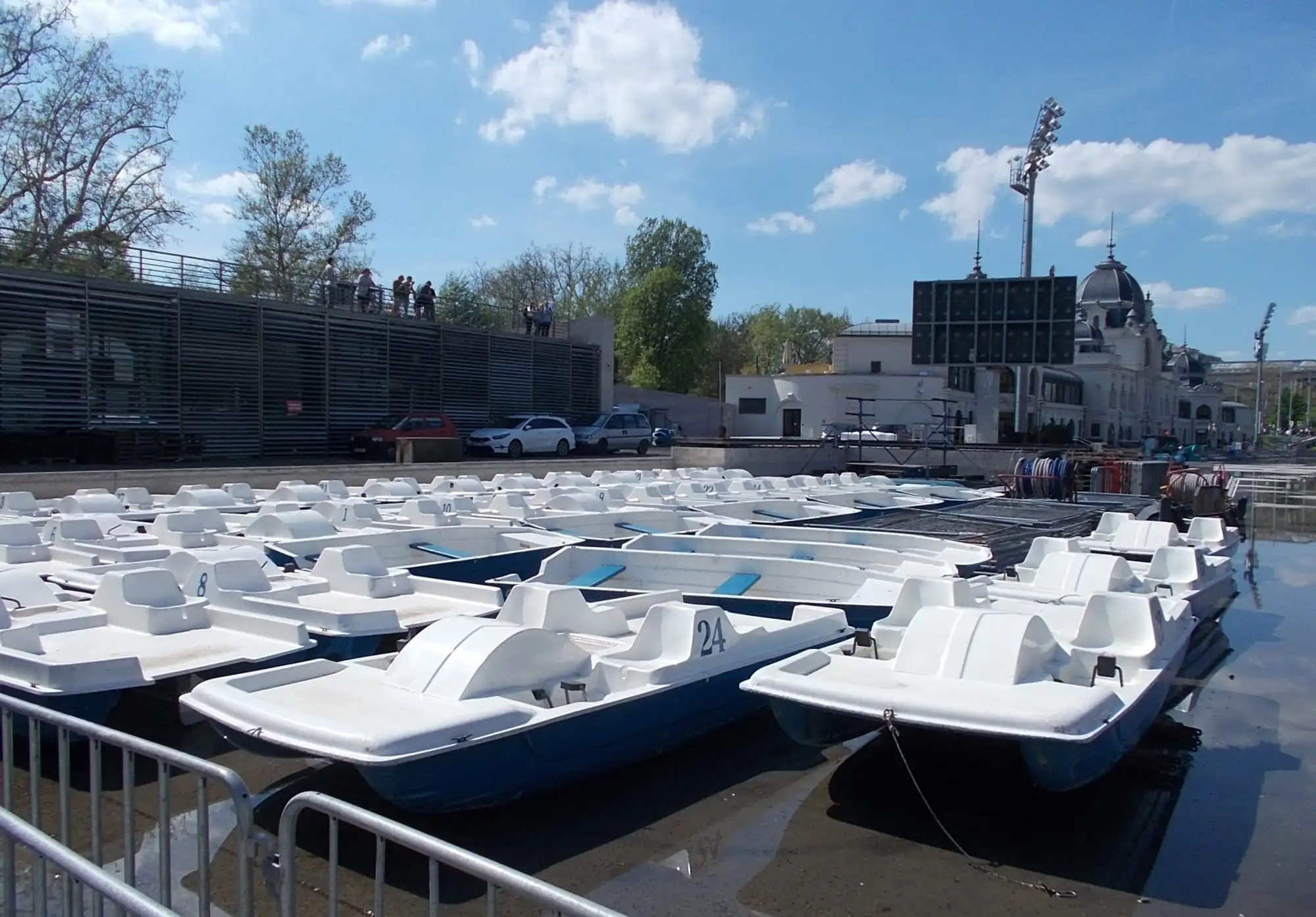Rows of white pedal boats moored at the edge of the City Park boating lake in Budapest, with the ice rink building behind them
