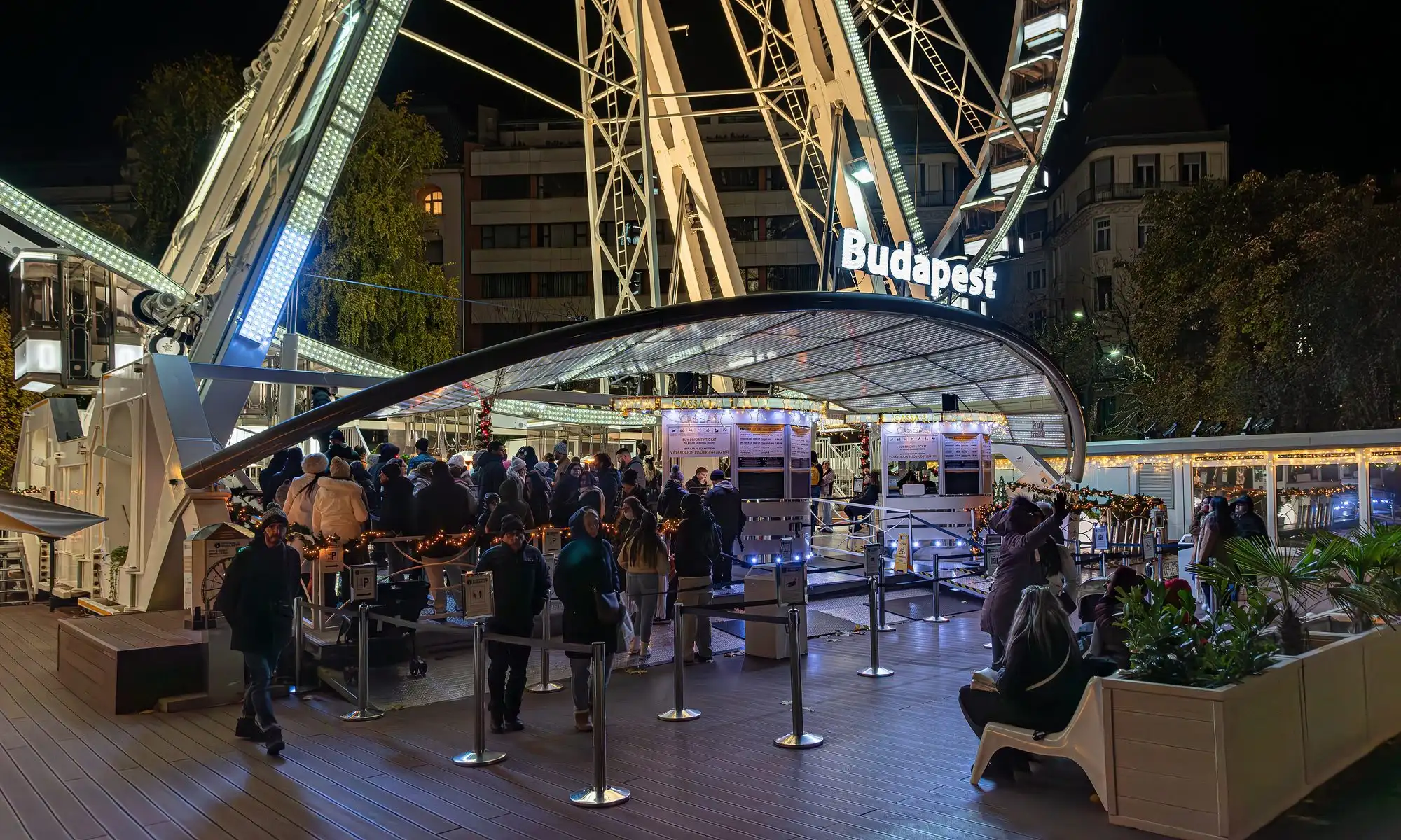 The lit boarding platform under the Budapest Eye at night, with a queue at the ticket windows and a cabin waiting at the left