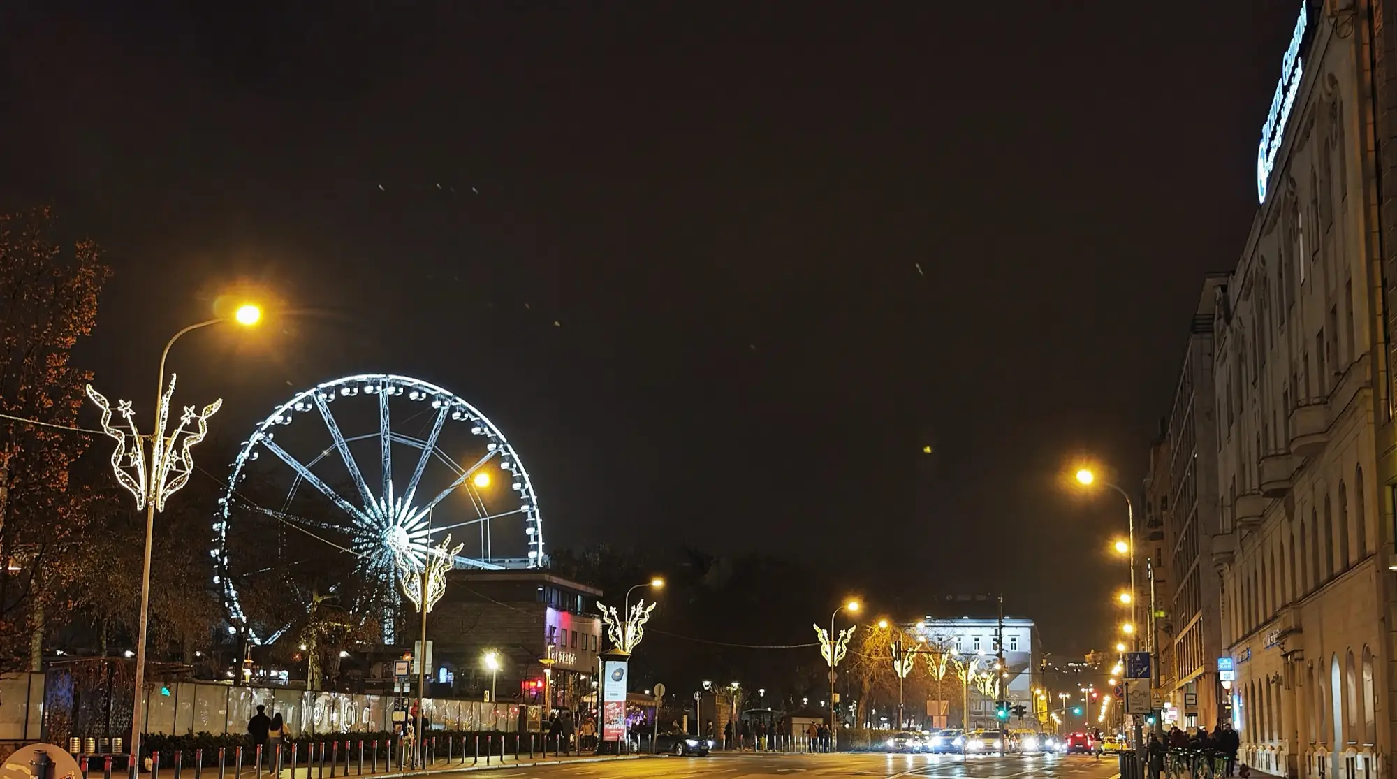 The Budapest Eye lit white at night beside a wide street hung with Christmas lights, with a few people on the pavement