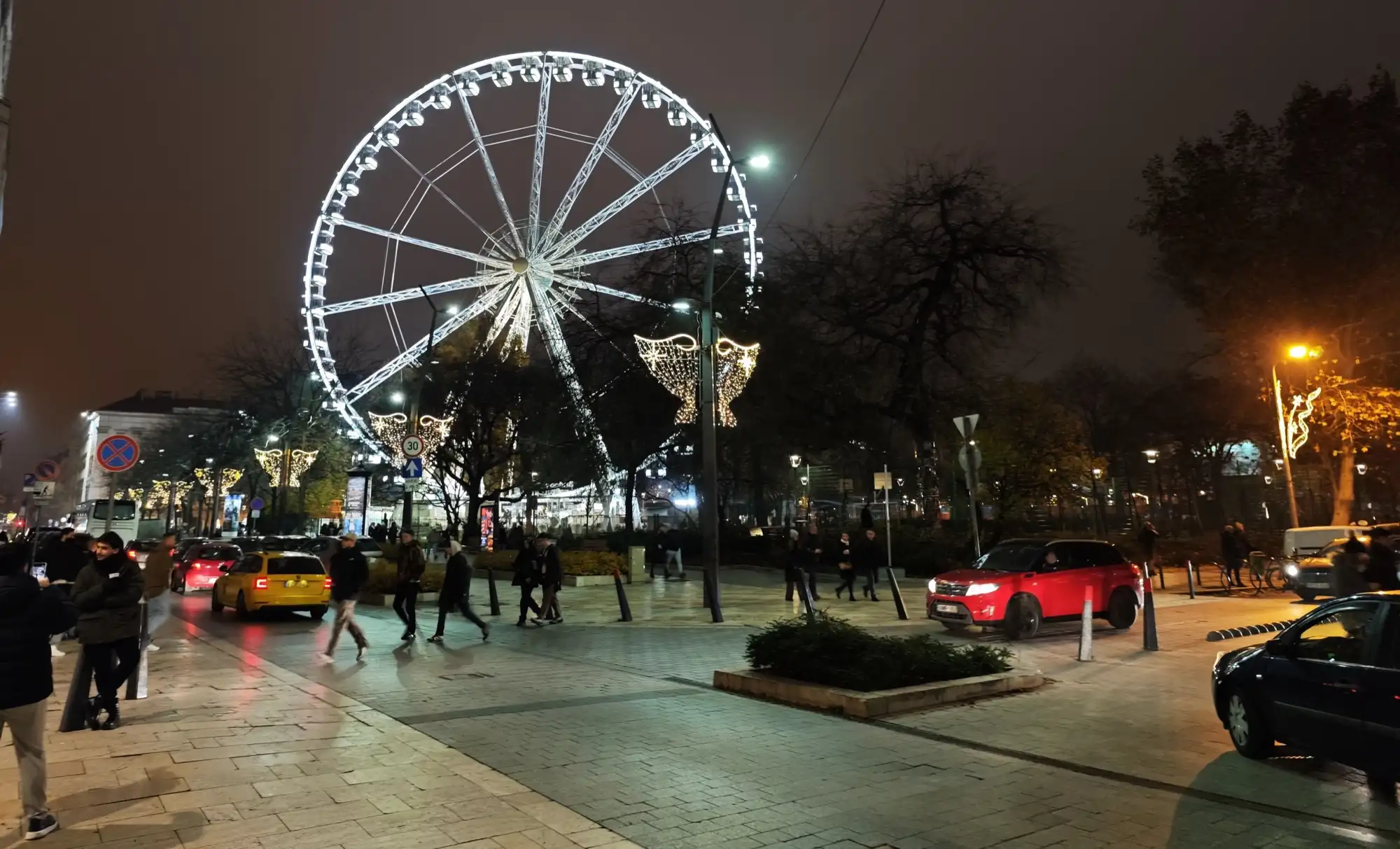 The Budapest Eye lit white at night on Erzsébet tér in December, with Christmas lights on the lamp posts along the street in front