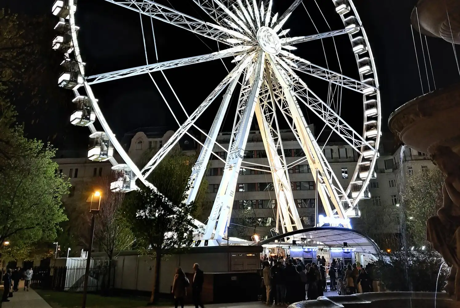 The Budapest Eye lit white at night over Erzsébet tér, with a queue at the ticket kiosk under its curved canopy and the Danubius Fountain at the right