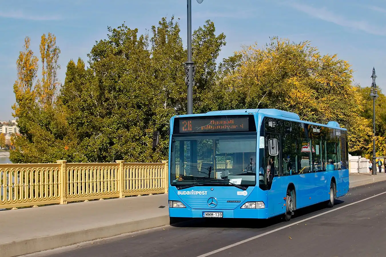 A blue BKK bus on route 26 to Nyugati pályaudvar, passing the yellow railings of the ramp onto Margaret Island with trees behind
