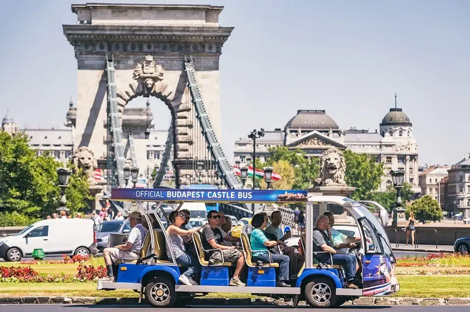 The blue Official Budapest Castle Bus electric minibus with passengers at Clark Ádám tér, with the Chain Bridge behind