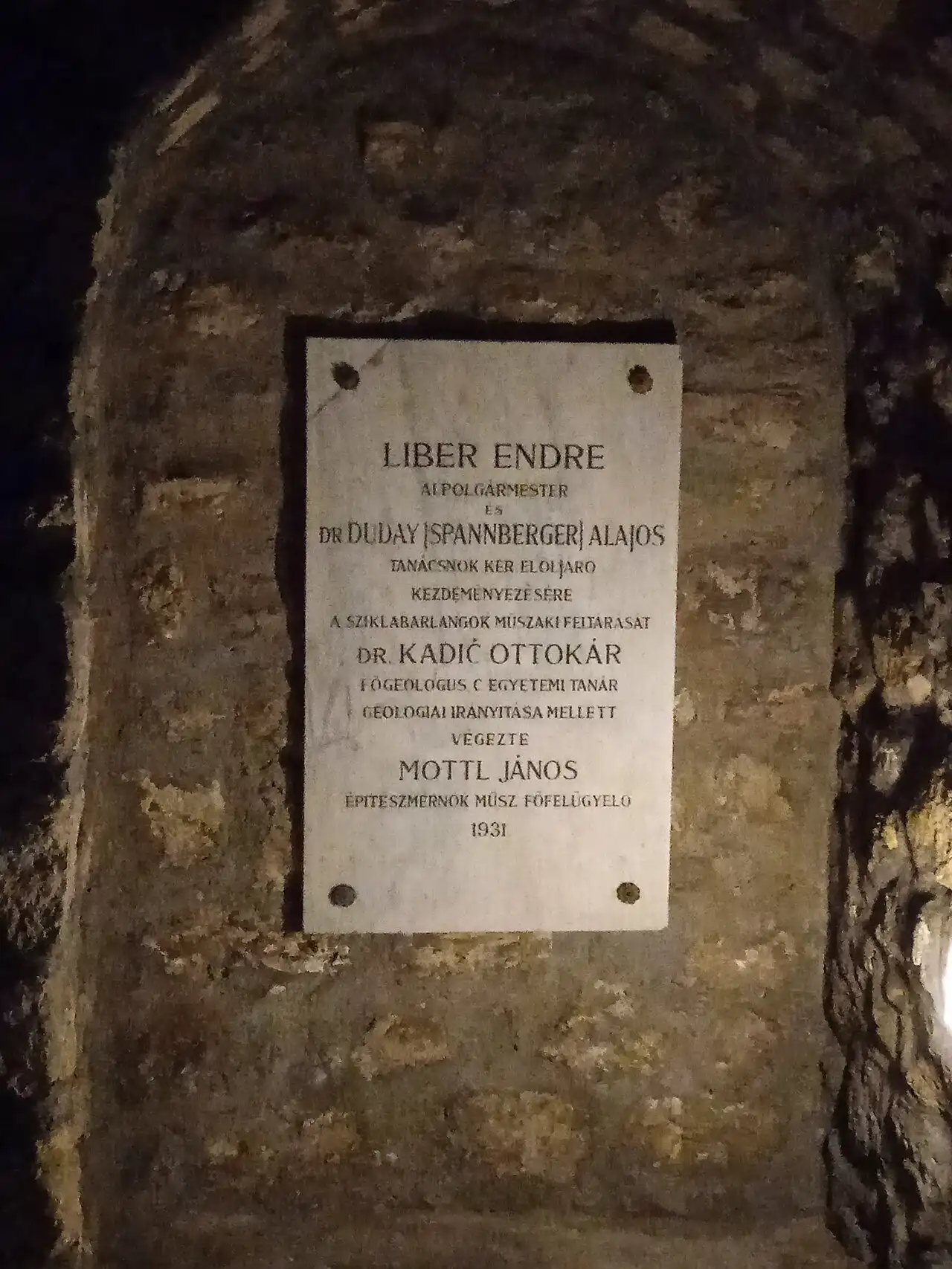 A white marble plaque set into the cave wall, dated 1931, recording the technical exploration of the rock cellars under the geological direction of Ottokár Kadić