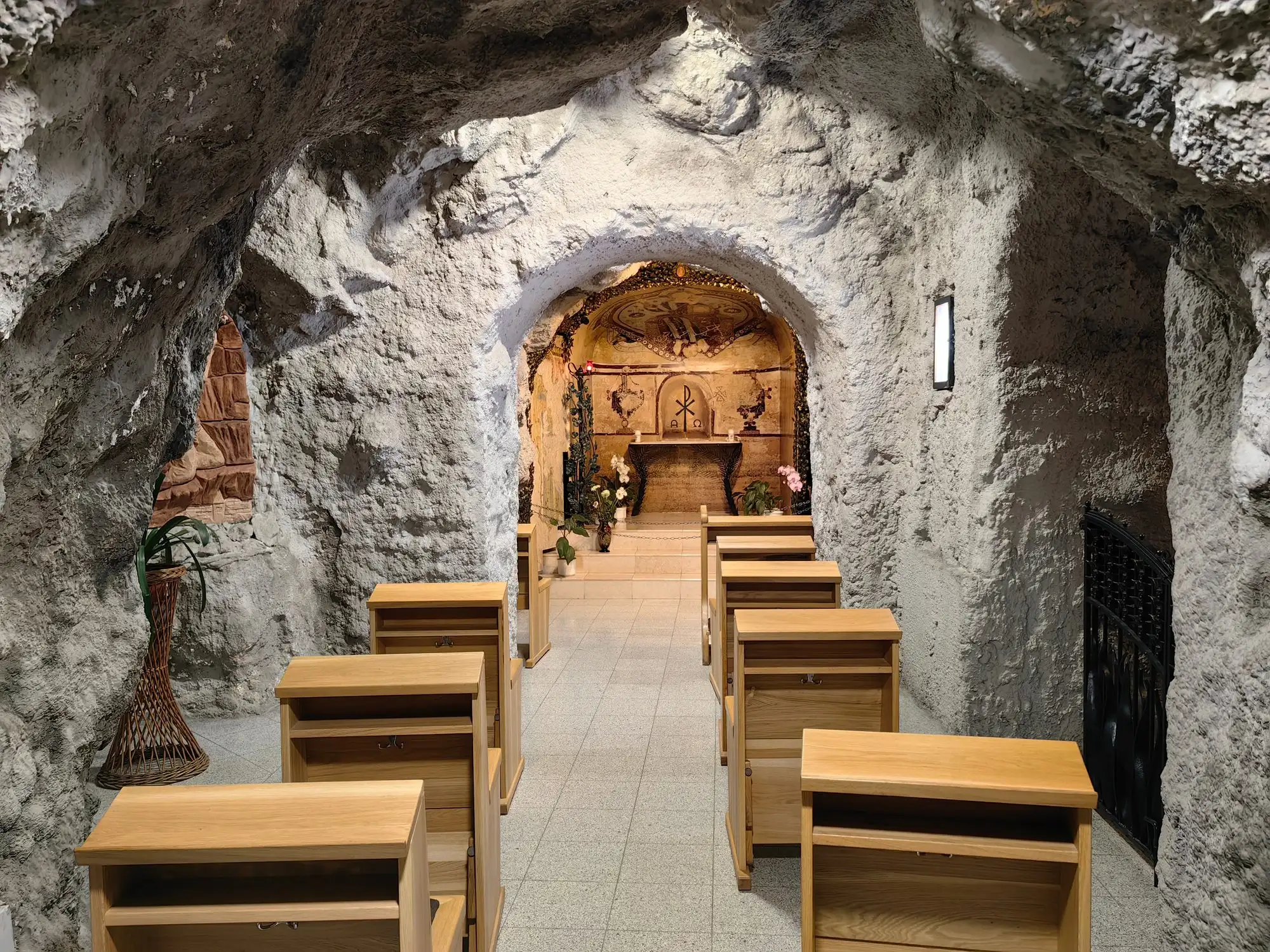 A narrow passage of the Cave Church cut through pale rock, with rows of wooden prayer desks leading to a small chapel with a carved wooden altar at the far end