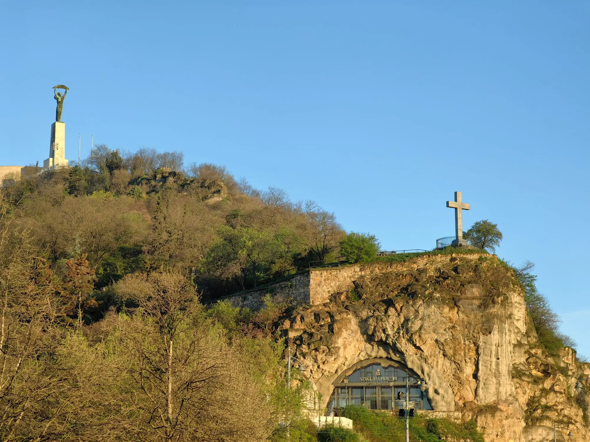 The glazed entrance of the Cave Church set into the rock at the foot of Gellért Hill, with a white cross on the cliff above it and the Liberty Statue on the summit behind
