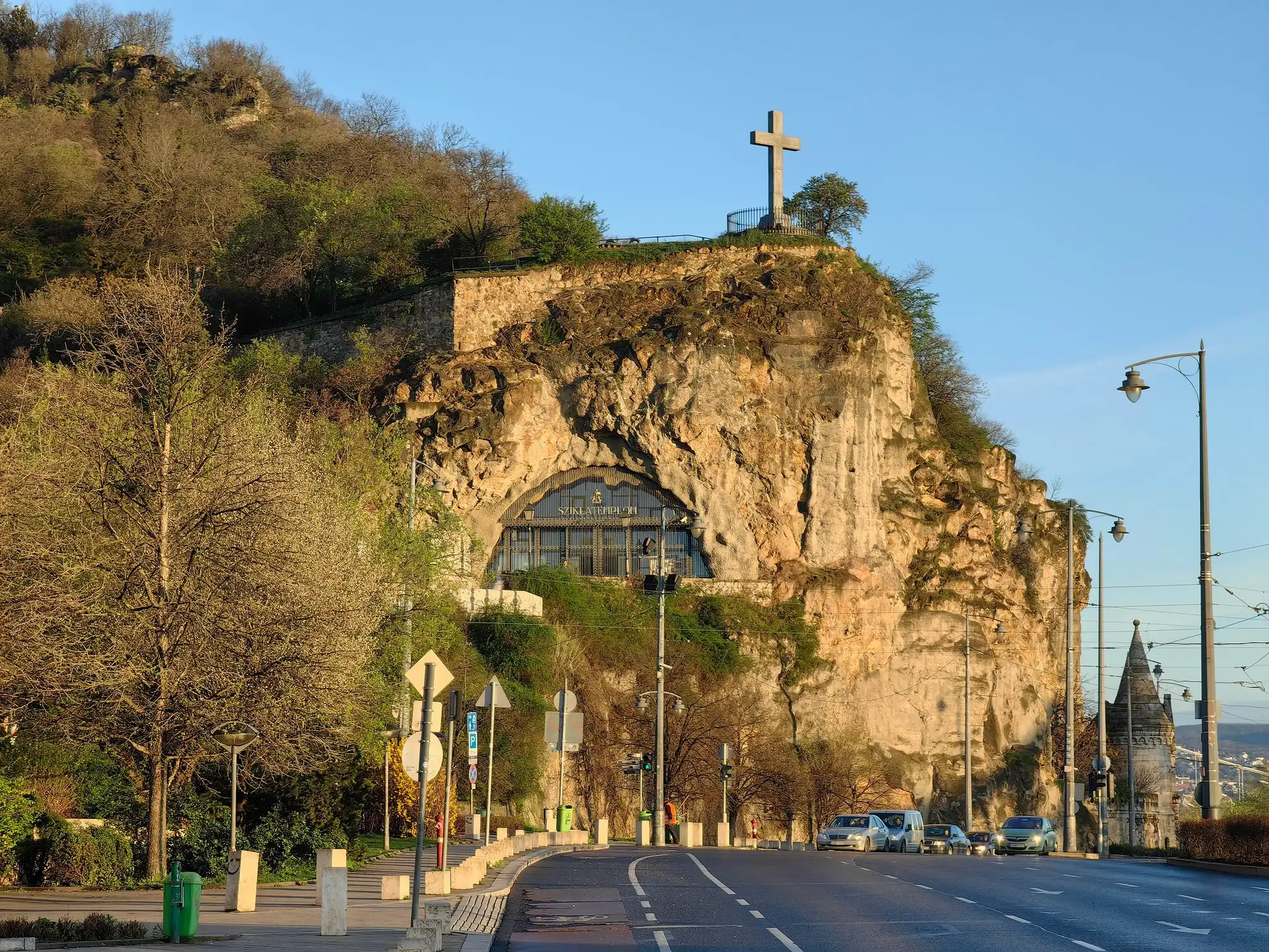 The arched entrance of the Cave Church set into the rock face of Gellért Hill above the embankment road, with a cross on the cliff above it