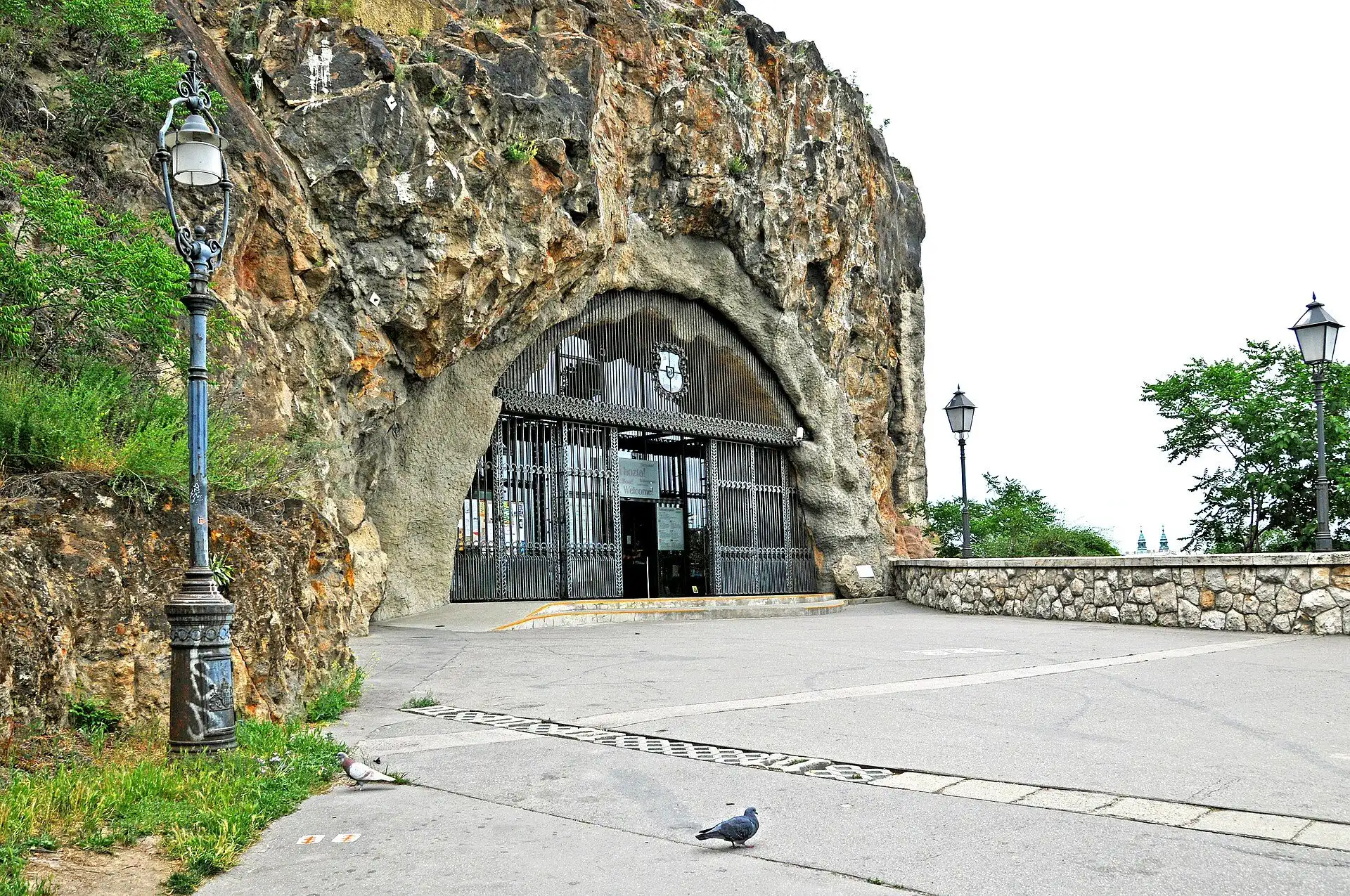 The arched mouth of the Cave Church in the rock face of Gellért Hill, closed by tall wrought-iron gates, with a paved terrace and street lamps in front