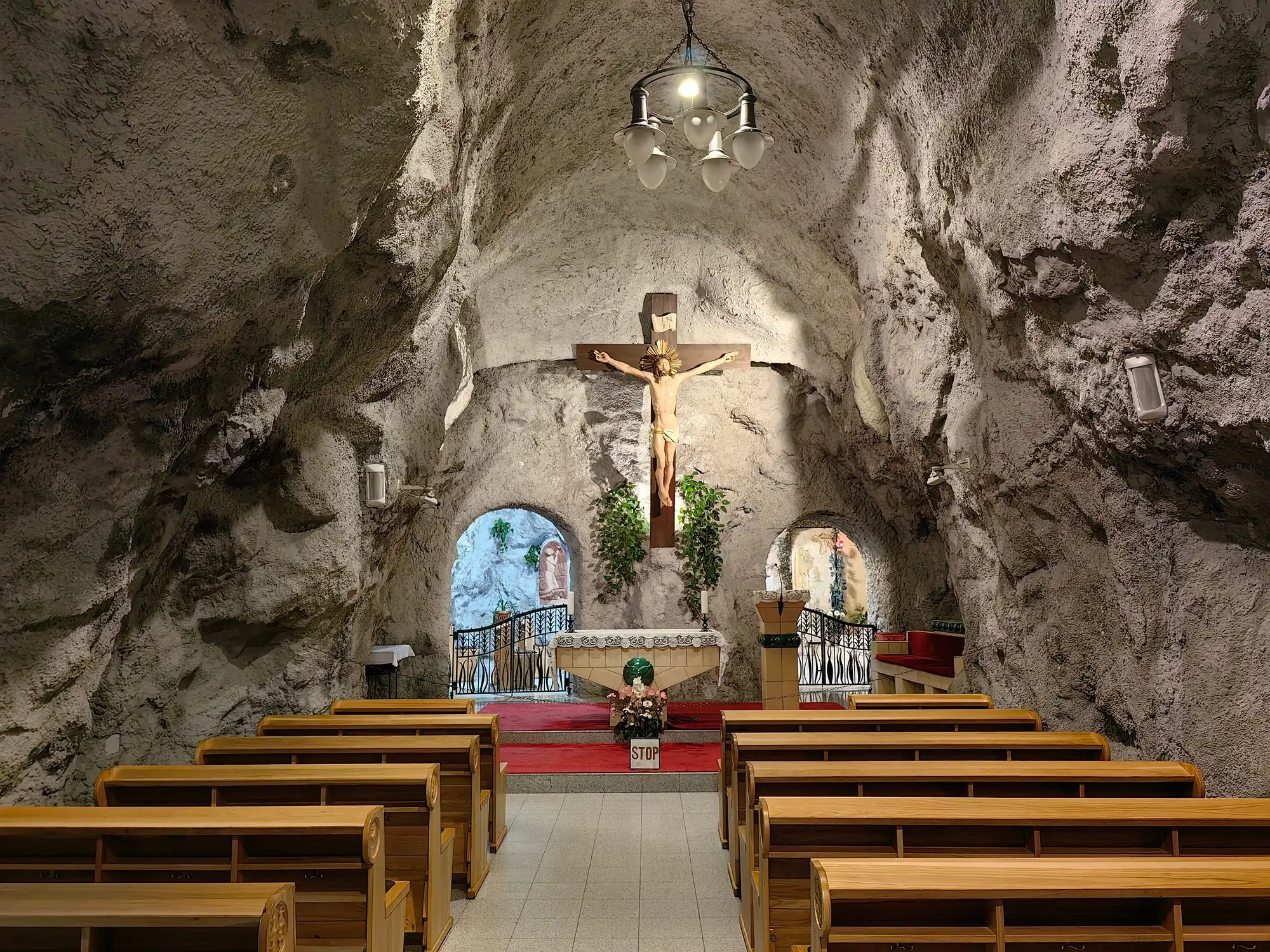 The main chapel of the Cave Church, rows of wooden pews facing an altar under a large crucifix, with rough rock walls and ceiling all around