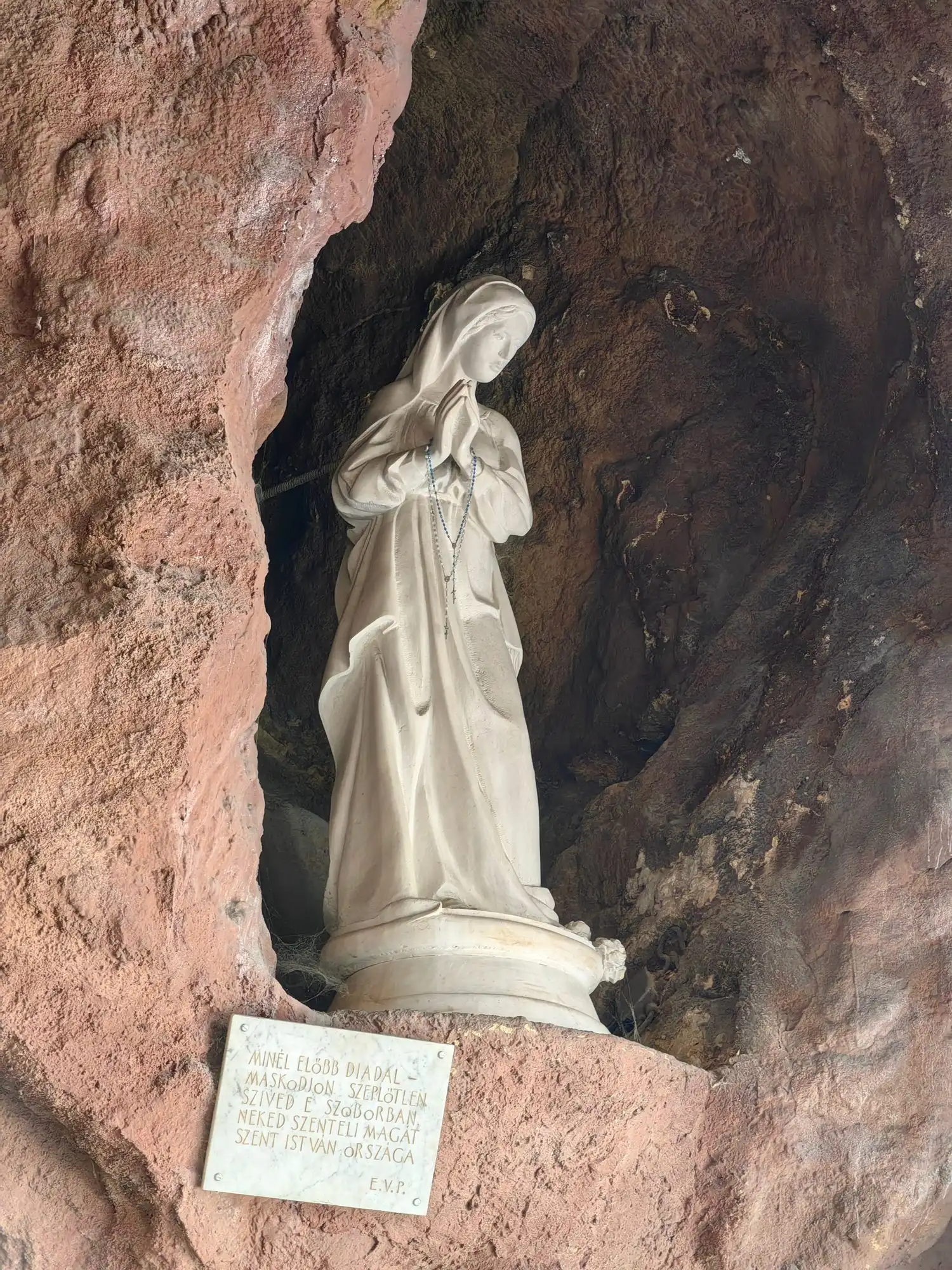 A white statue of Our Lady of Lourdes, hands joined in prayer, standing in a niche in the reddish rock of the outer cave, with a small plaque below
