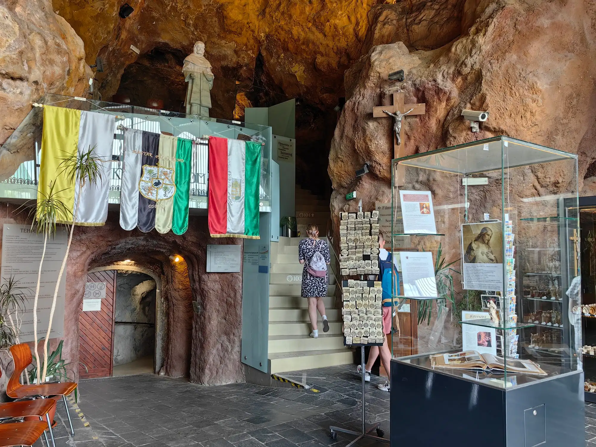 The outer cave of the Cave Church, with a gift shop counter and display cases, a row of flags hanging from the rock and steps leading up to a door into the church