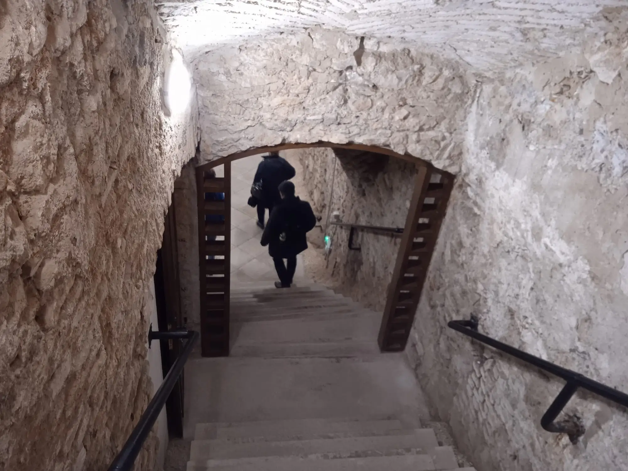 A lit stone stairway leading down through a doorway into the Buda Castle Cave, with a handrail on one side