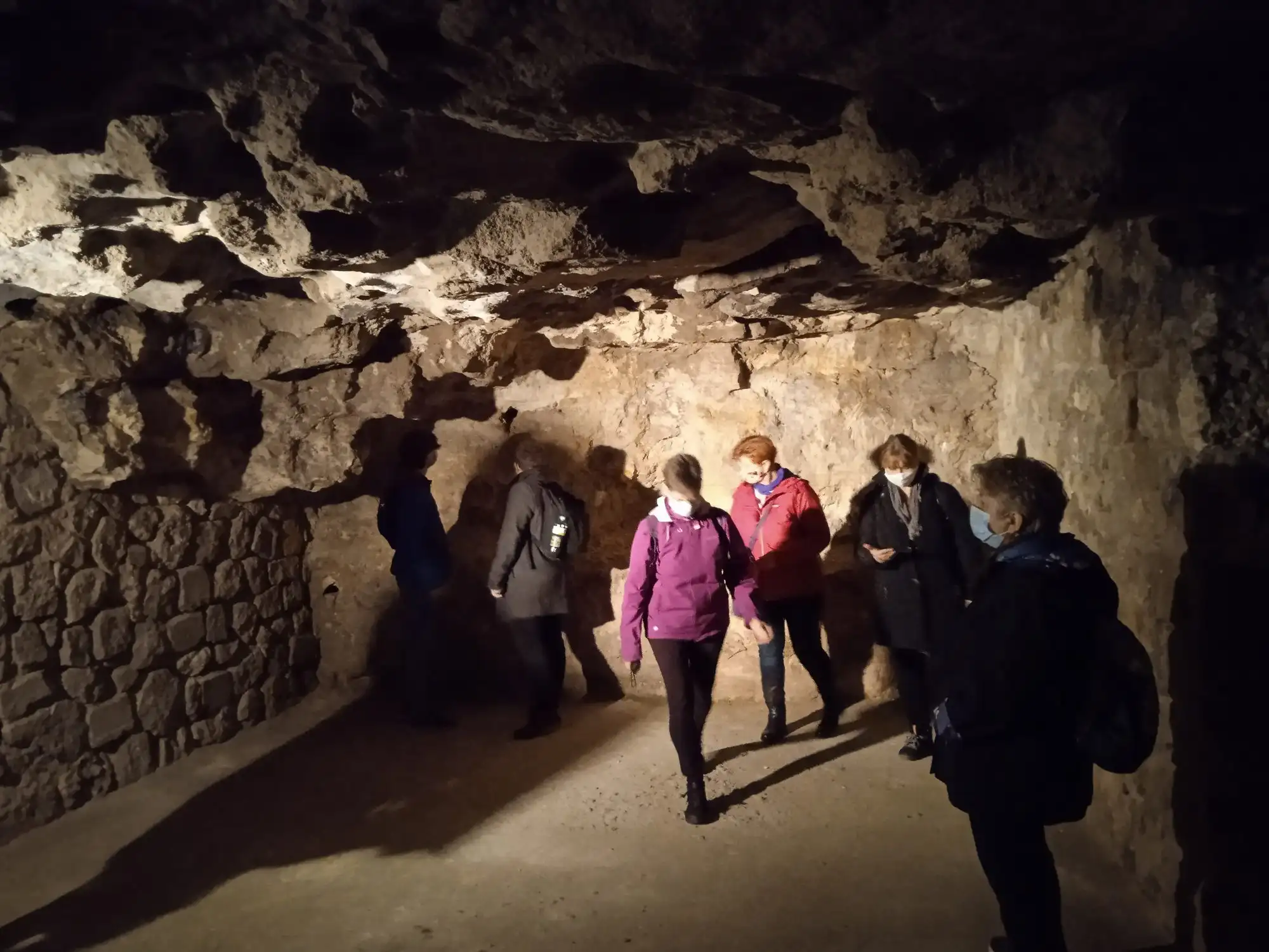 A guided group walking through a chamber of the Buda Castle Cave, under a natural rock ceiling above a dry-stone wall