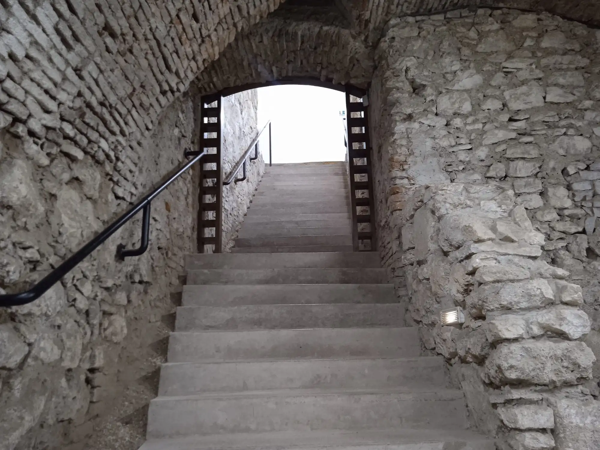 A lit stone staircase leading up out of the Buda Castle Cave towards daylight, with a handrail on one side