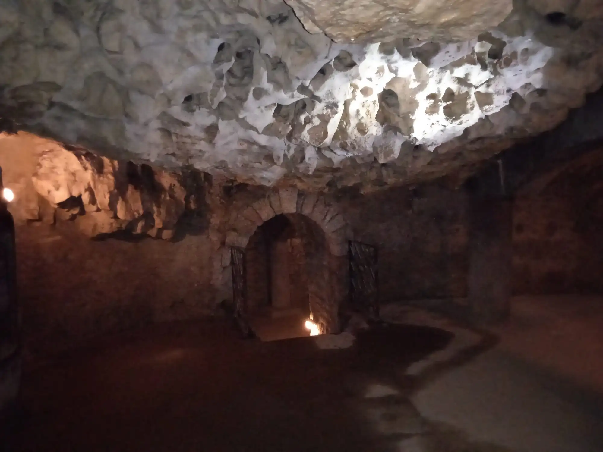 A pitted white travertine ceiling above a cut stone chamber with an arched, gated doorway in the Buda Castle Cave