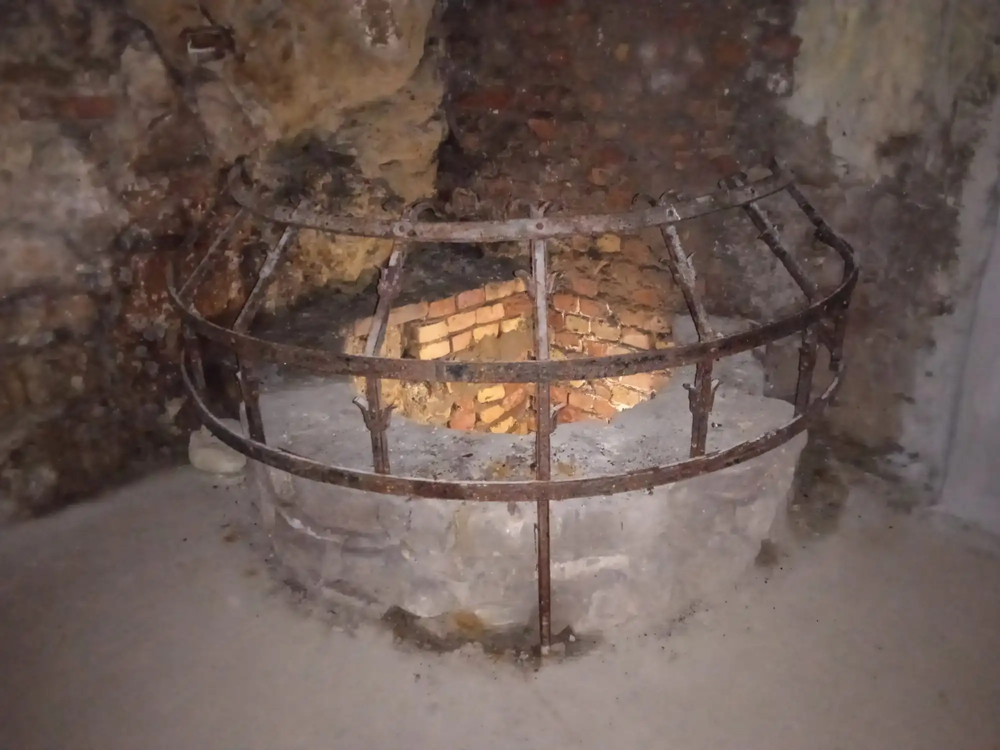 A round medieval well head with a rusted iron railing, lit from inside, in a chamber of the Buda Castle Cave