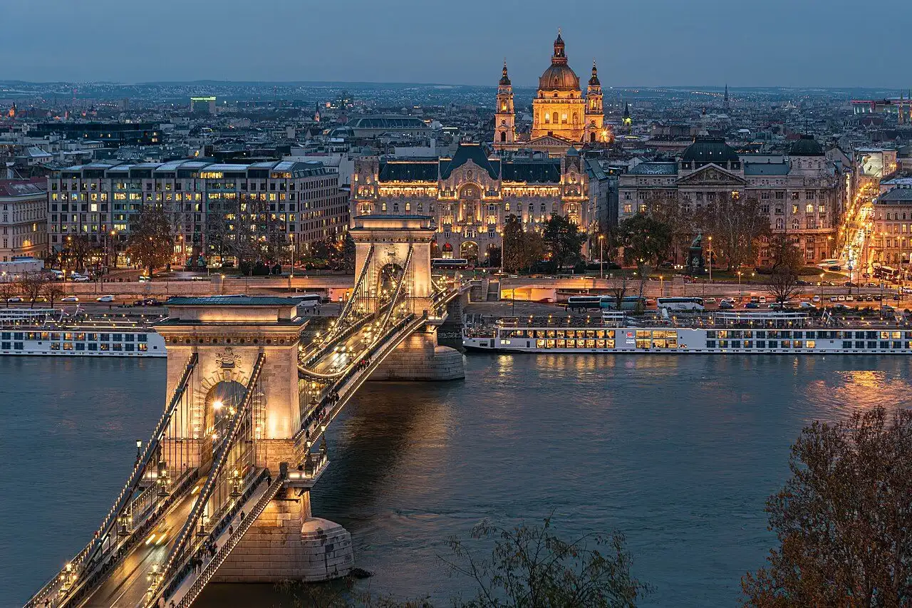 The Széchenyi Chain Bridge lit at dusk, its chains picked out in warm white light, with the Gresham Palace and the dome of St Stephen's Basilica on the Pest bank behind