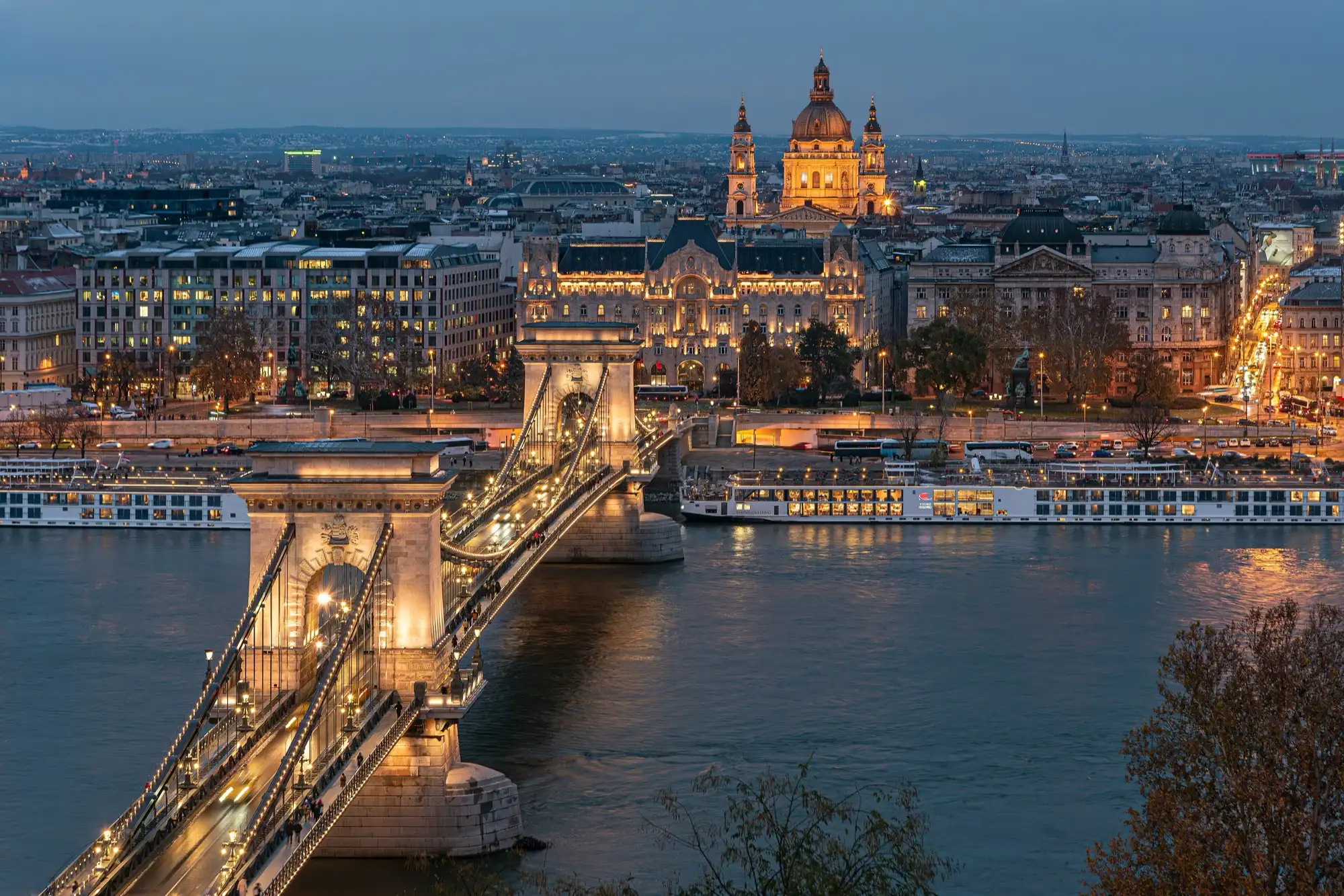 The Chain Bridge lit at blue hour over the Danube, seen from the Buda side, with Gresham Palace and the dome of St Stephen's Basilica lit up behind it