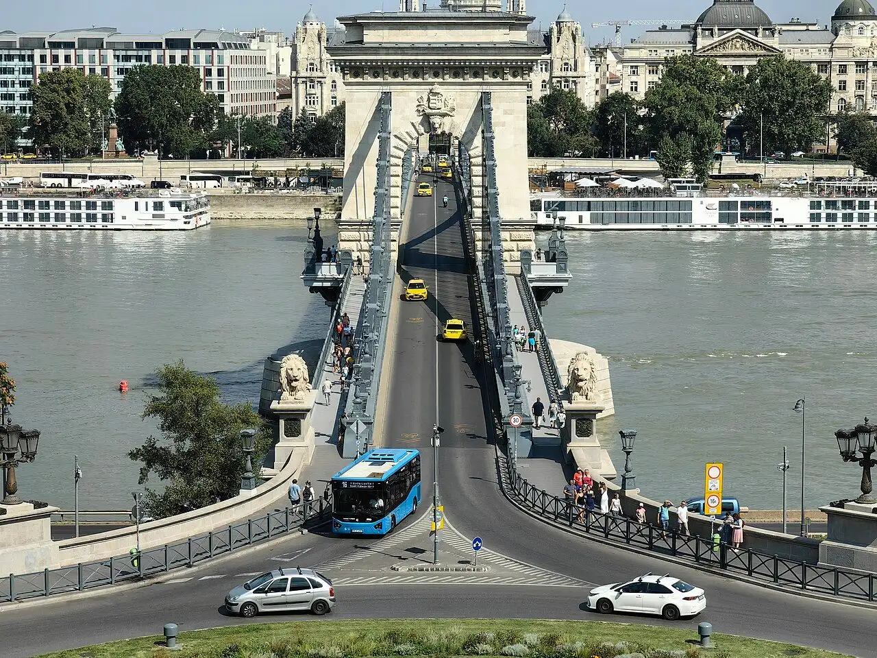 A blue BKK bus turning onto the Chain Bridge at the Buda end, with taxis on the roadway and cars circling the Clark Ádám tér roundabout