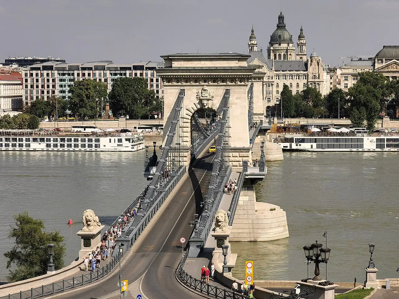 The Széchenyi Chain Bridge seen from Castle Hill, with pedestrians on both pavements, the two Buda lions in the foreground and the dome of St Stephen's Basilica beyond