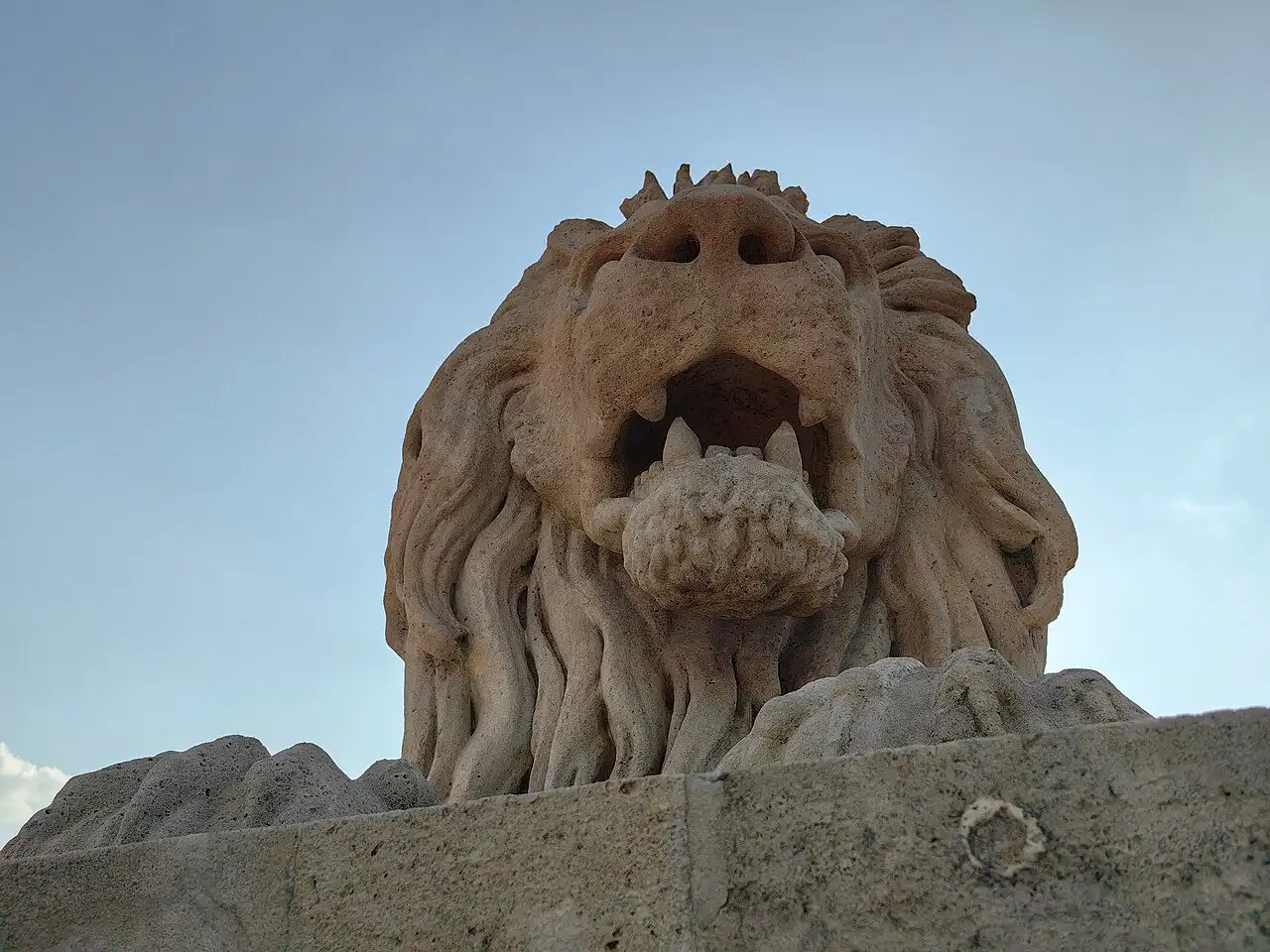 The head of one of the Chain Bridge lions seen from below, its jaws open and the tongue visible inside the mouth