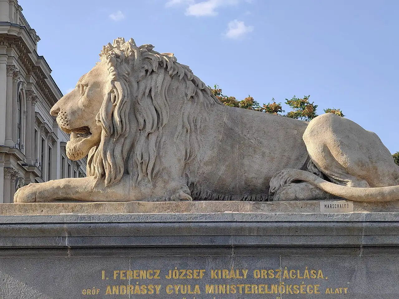 A Chain Bridge lion on its plinth, with the sculptor's name carved on the plinth edge and a gilded inscription along the side below it