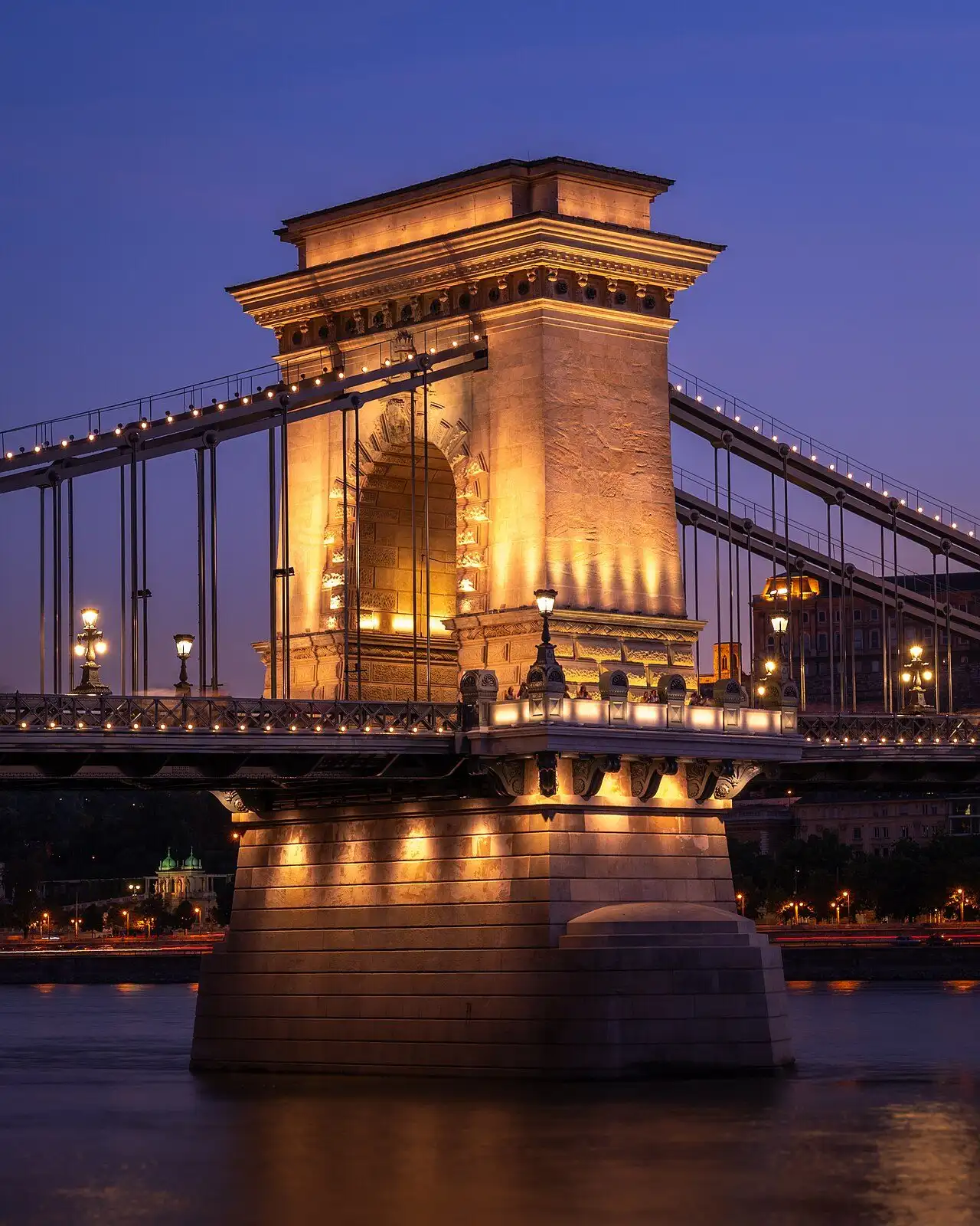 A river pier of the restored Chain Bridge lit at dusk, showing the gateway arch, the chains, the lamps and the pavement running round the outside of the stonework