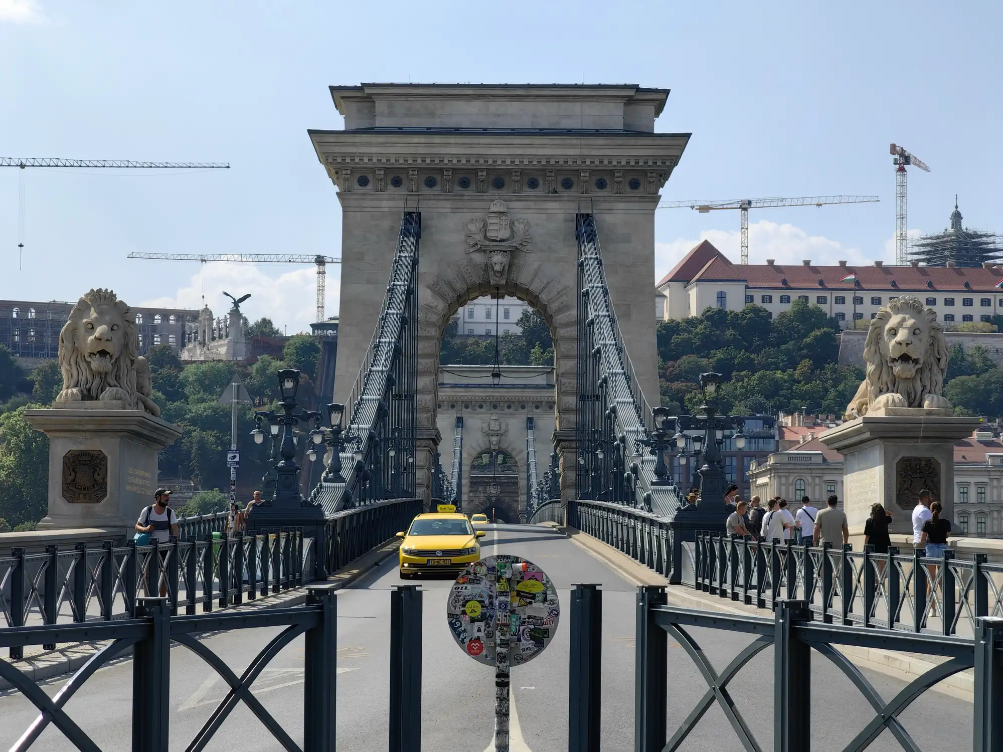 A yellow taxi driving off the Chain Bridge at the Pest end between the two lions, with people walking on the pavements either side and Castle Hill behind the river pier