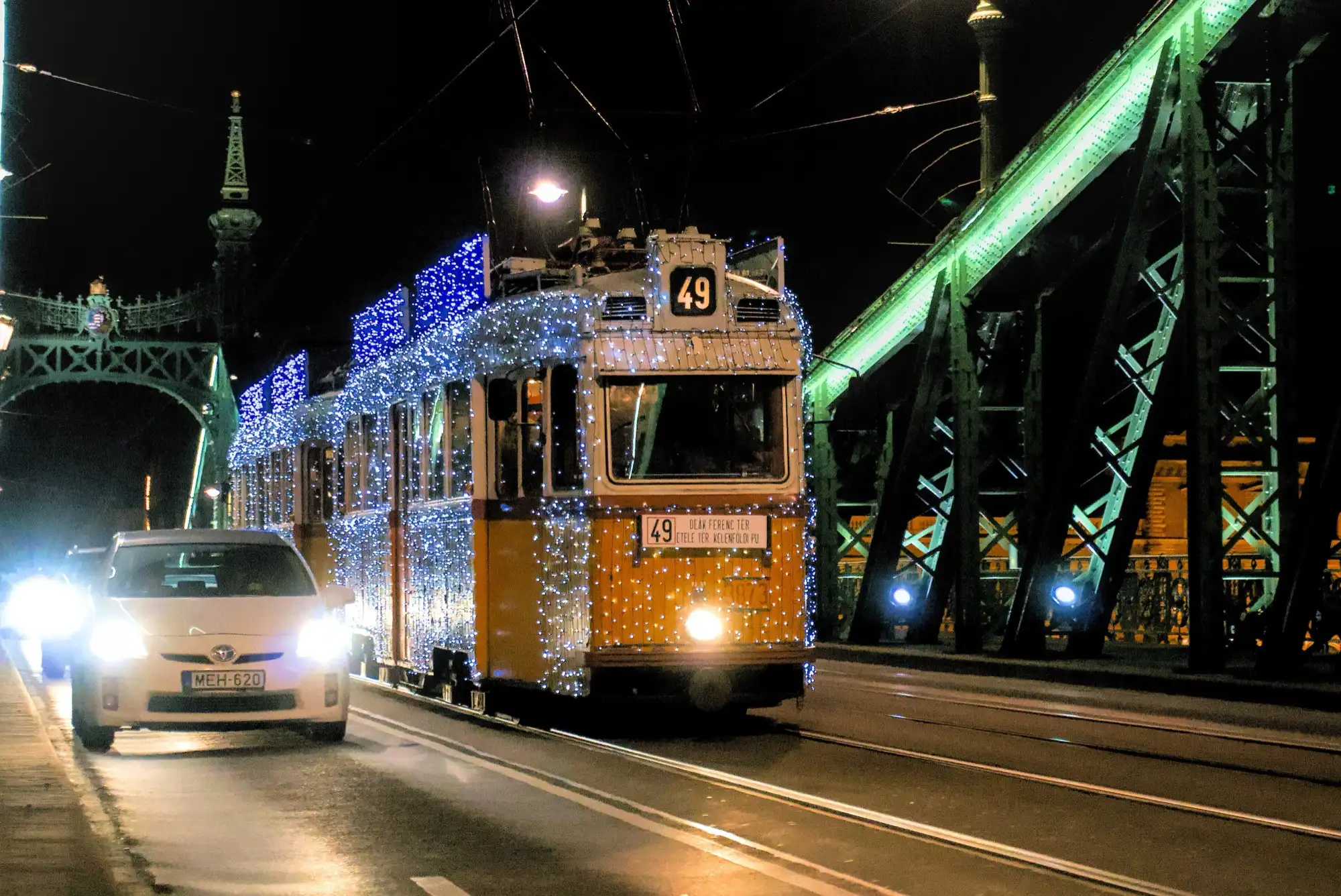 A yellow Budapest tram covered in blue and white Christmas lights crossing Liberty Bridge at night