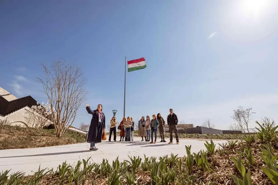 A guide and a small group standing on the paved walk in the Citadella's new park, beside the tall mast flying the Hungarian flag