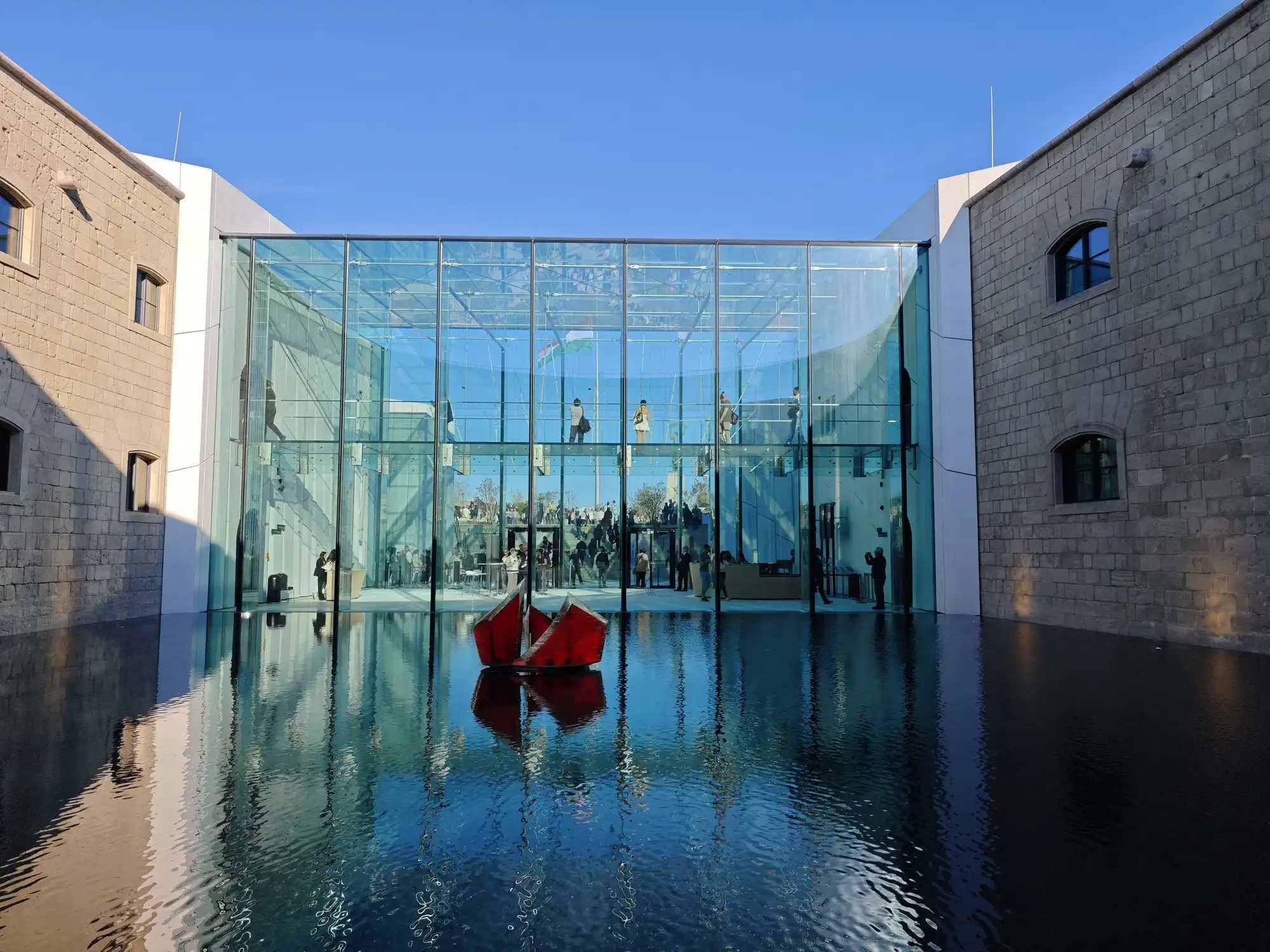 The reflecting pool in the former round-bastion courtyard of the Citadella, a red sculpture at its centre and the glass hall behind it mirrored in the water between the old stone walls