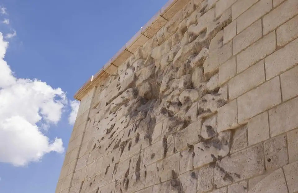 A stretch of the Citadella's pale stone fortress wall with a patch of stonework torn and pitted by explosive fragments, against a blue sky