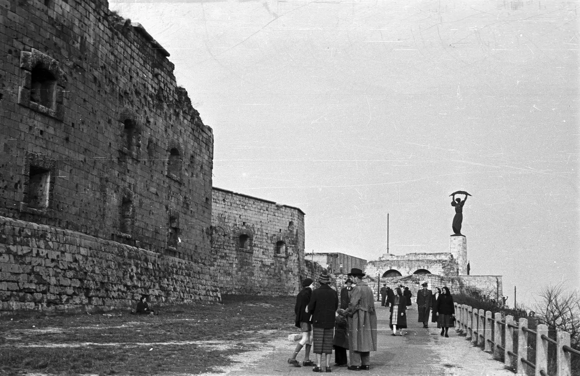 The rough stone wall of the Citadella in 1953, with people walking the promenade beside it and the Liberty Statue standing beyond