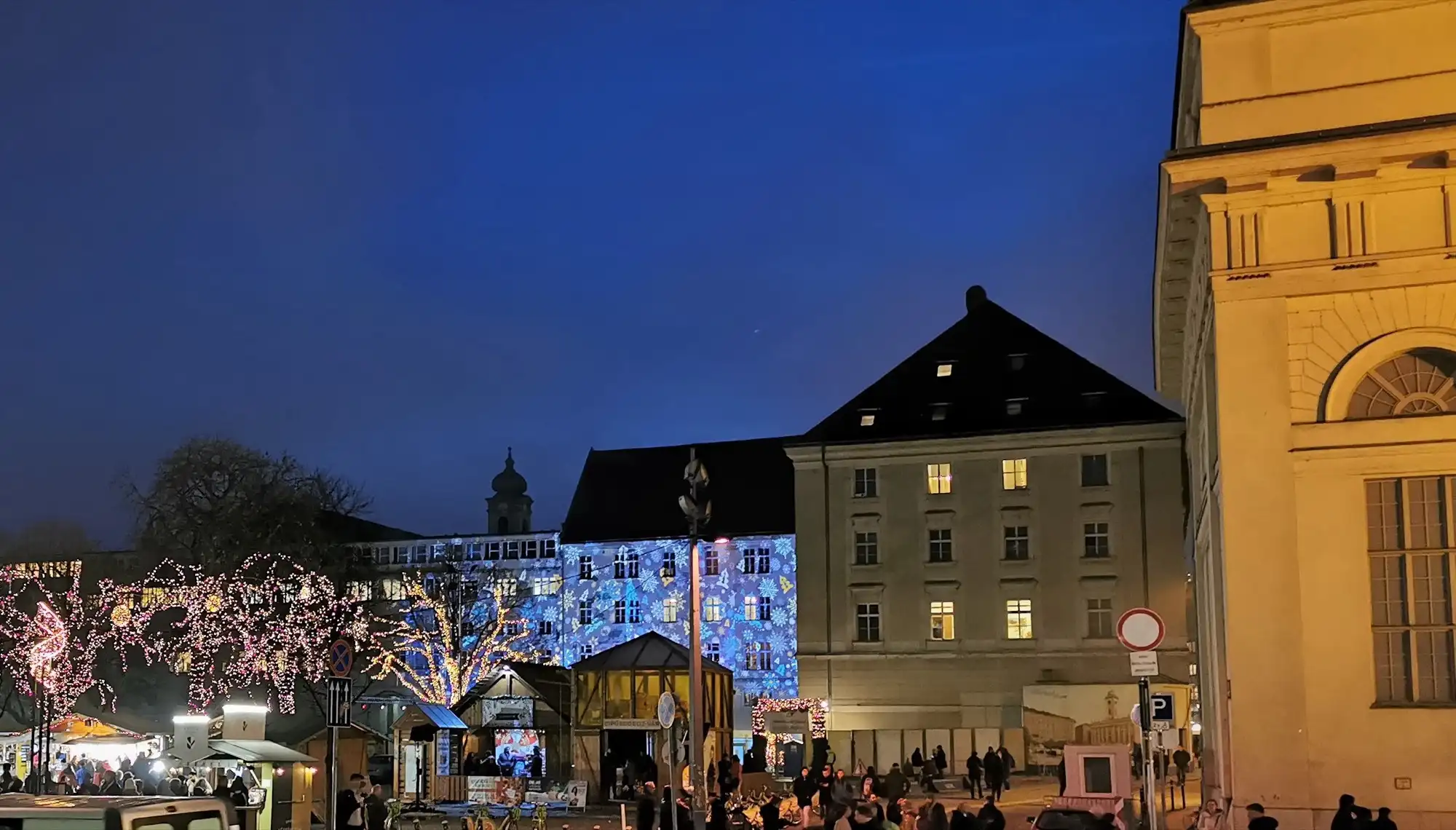 Snowflakes projected in blue light across a wing of the Budapest City Hall at dusk in a past season, with trees wrapped in pink and gold lights in front