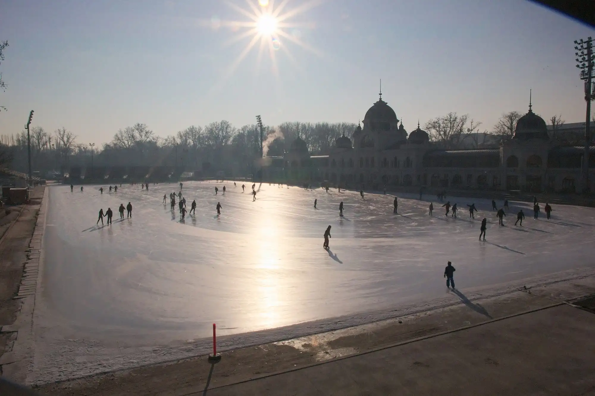 Skaters spread across the wide outdoor ice of the City Park Ice Rink in Budapest at low winter sun, with the rink's domed building behind