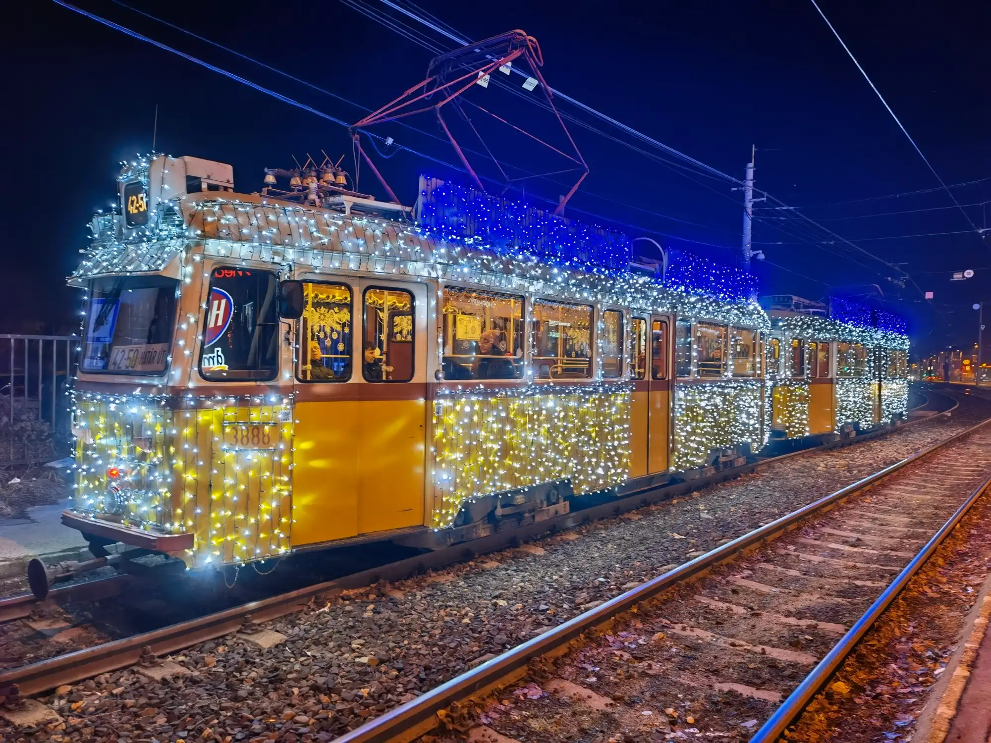 The classic Budapest Christmas tram, an old yellow tram covered from end to end in white, gold and blue lights, at a stop at night