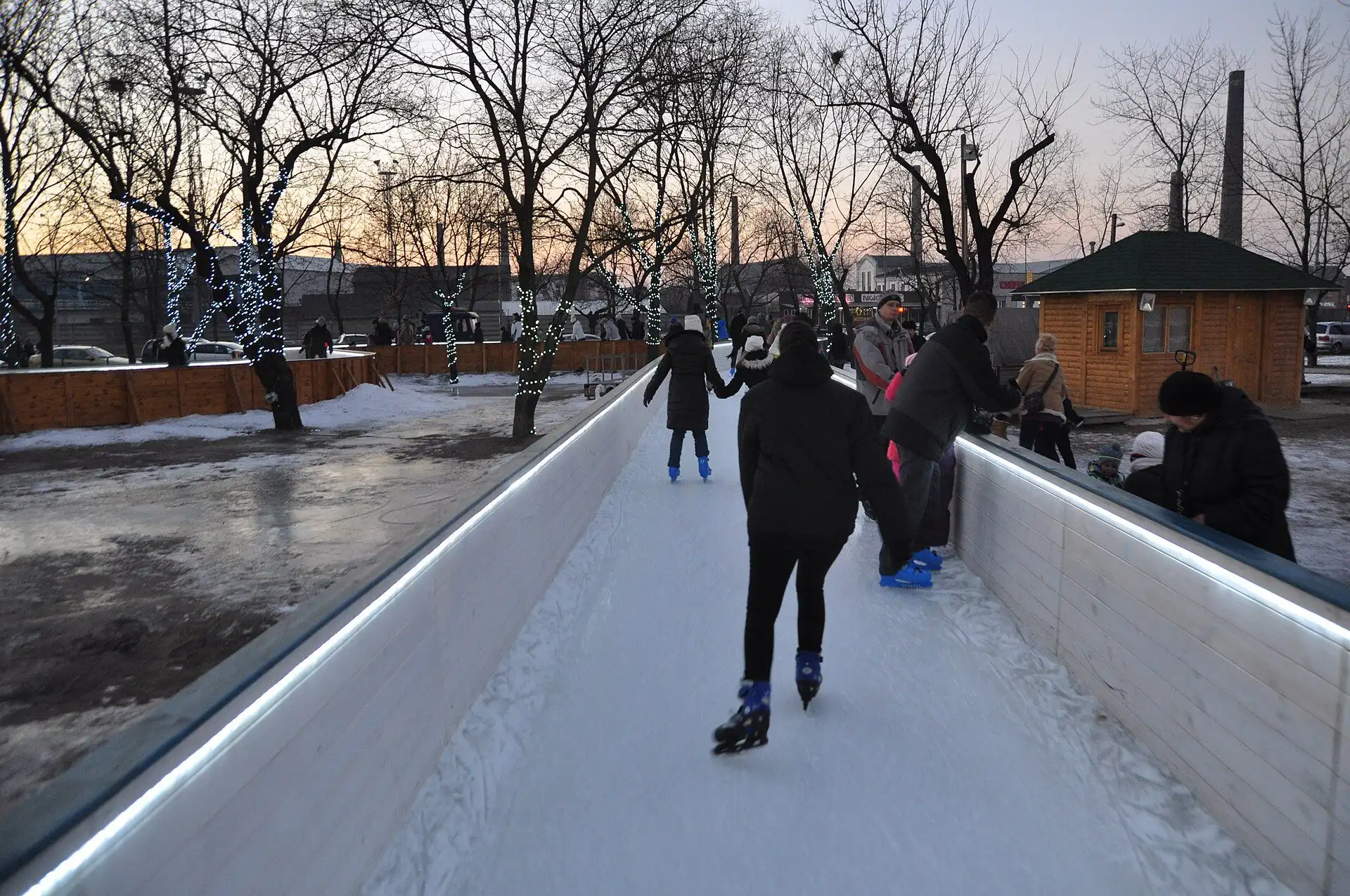 Skaters on a narrow ice corridor between low white boards and lit trees at the Csepel ice rink at dusk