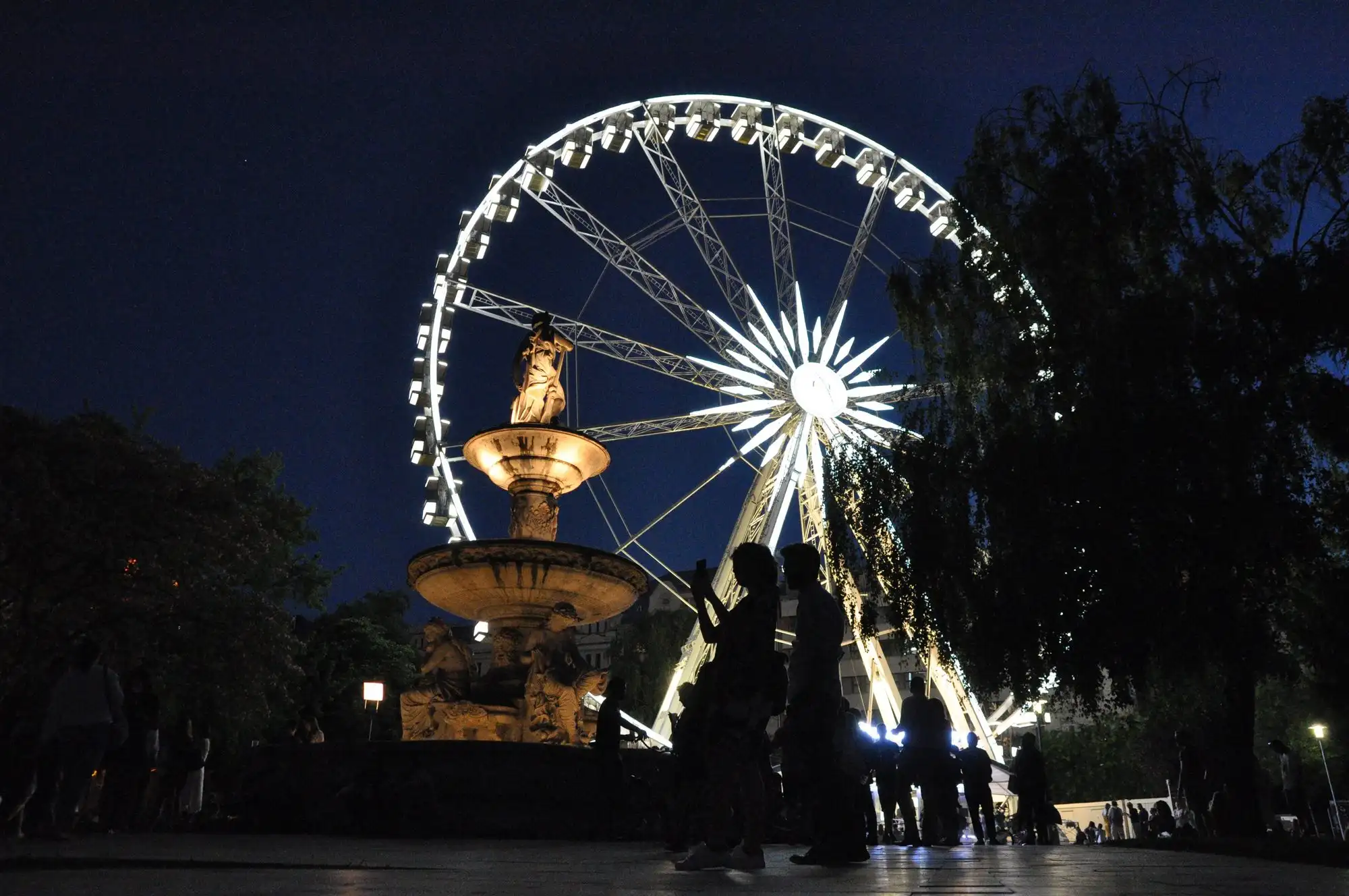 The Danubius Fountain lit up at night on Erzsébet tér with the illuminated Budapest Eye turning behind it